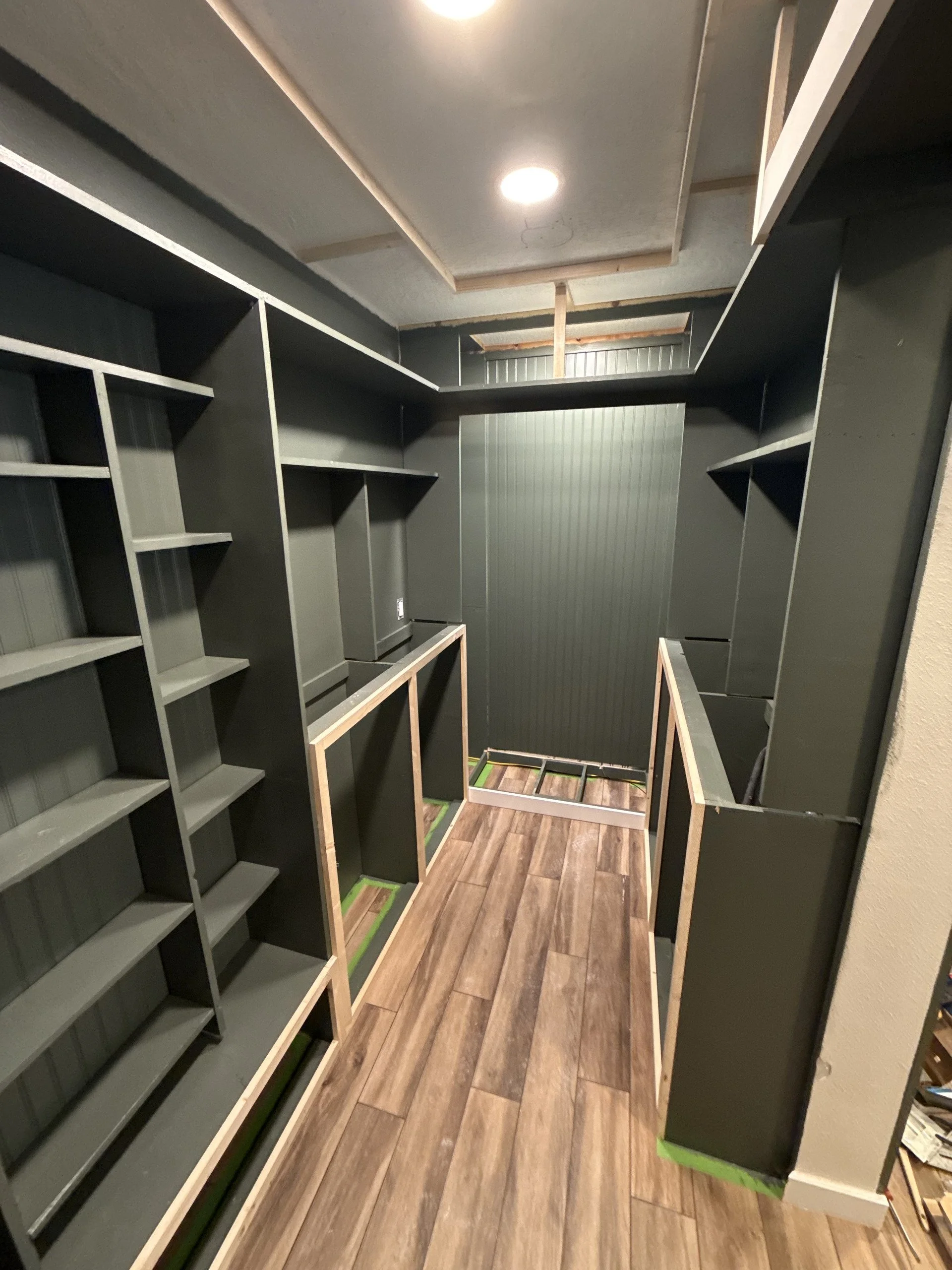 Photo of a closet under construction with black built-in shelves on the walls, a small framed opening at the base, wood plank flooring, and unfinished walls and ceiling.