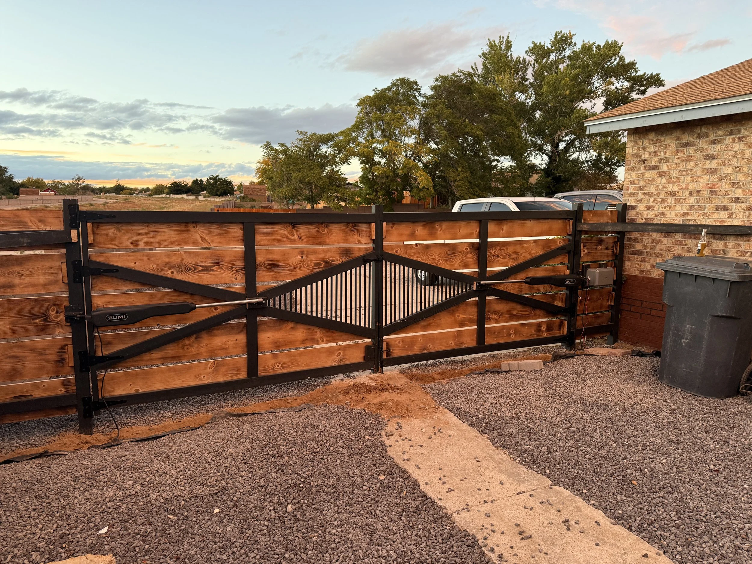 View of a backyard with a wooden and metal gate, a brick house, a trash bin, a tree with green and yellow leaves, and several parked cars under a cloudy sky at sunset.