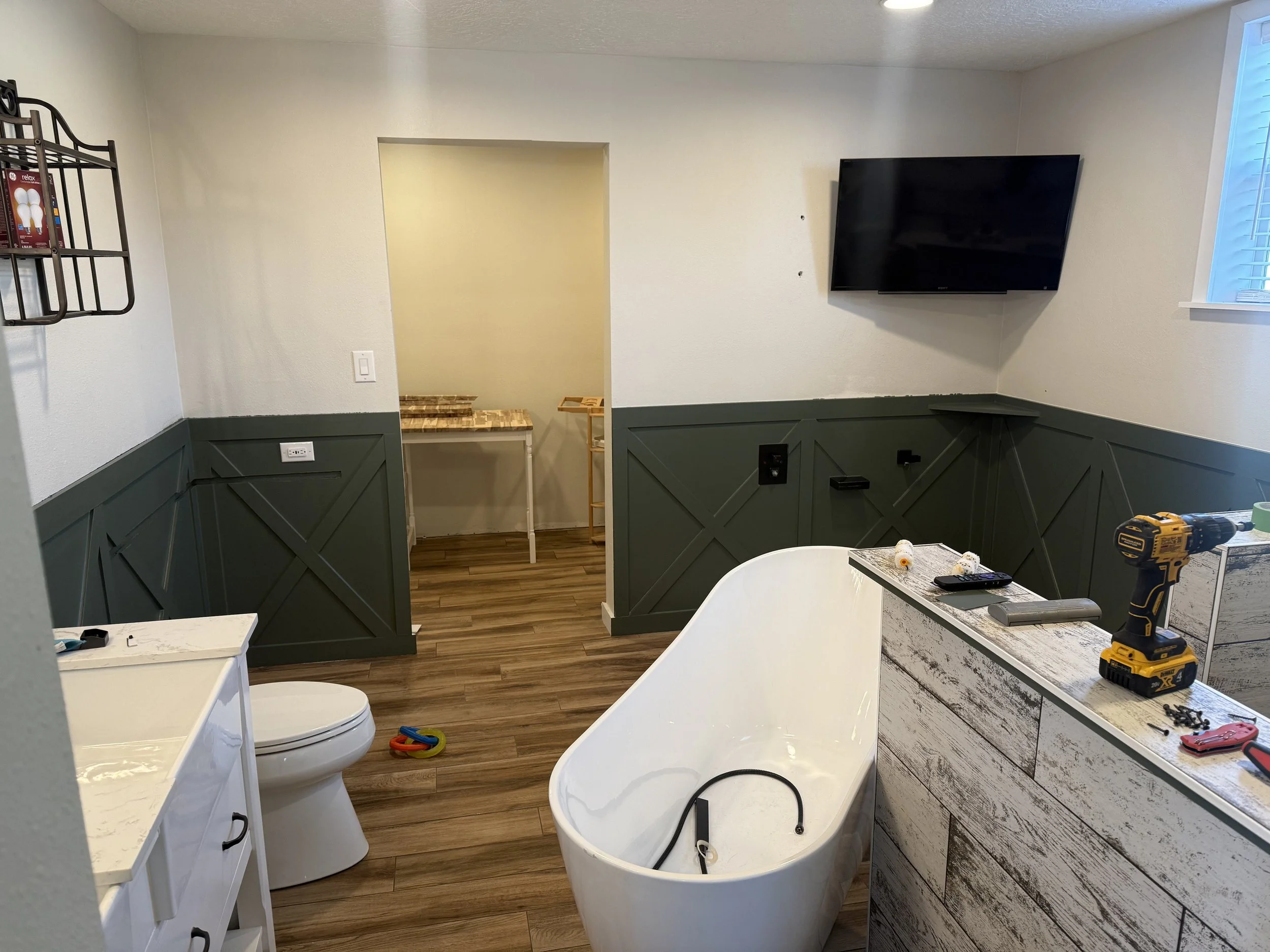 A room in the process of renovation with a partially installed bathtub, hanging tools, and construction materials. There is a wall-mounted TV on the right and a window with blue blinds on the far right. The lower part of the walls is painted dark gre