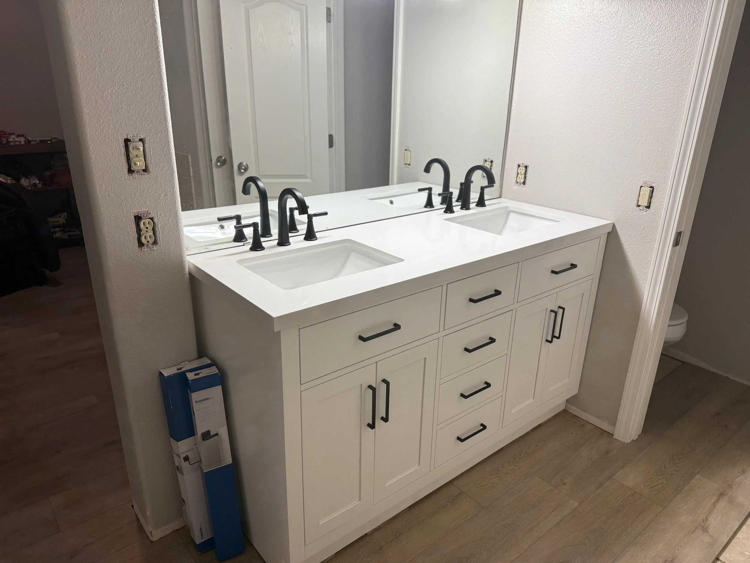 Bathroom vanity with two sinks, black faucet fixtures, white cabinet, and large mirror. Electrical outlets are visible on the wall, with some covers missing. The floor is wooden.