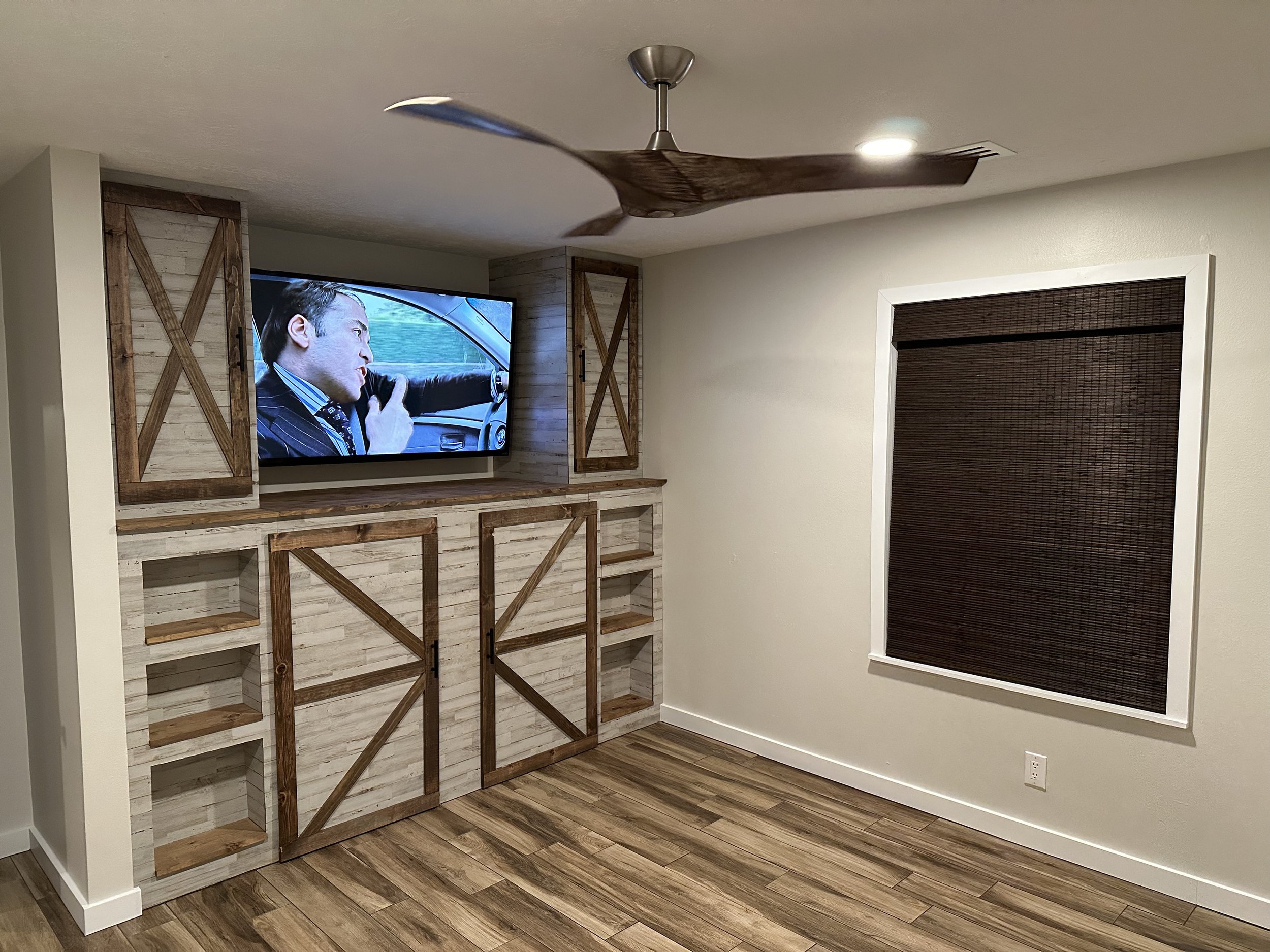 Bedroom featuring a ceiling fan, a flat-screen television mounted on the wall, and wooden shelving with a rustic finish. The walls are painted off-white and there is a window with dark brown blinds. The floor is wood-patterned.