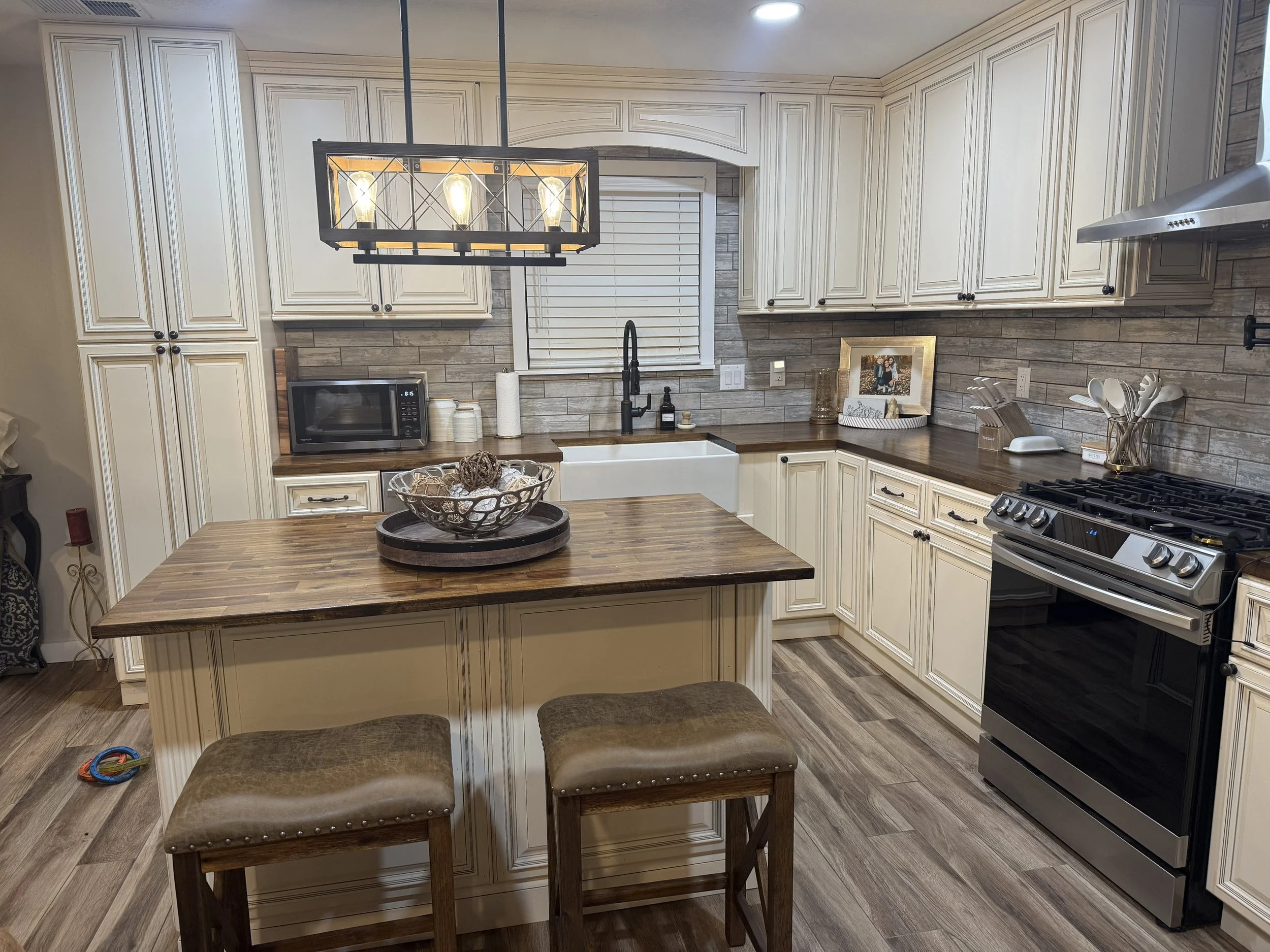 Modern kitchen with cream-colored cabinetry, wooden countertops, and a gray tiled backsplash. Features include a microwave, decorative items, a framed photo, and a stainless steel stove. A kitchen island with stools is in the foreground.