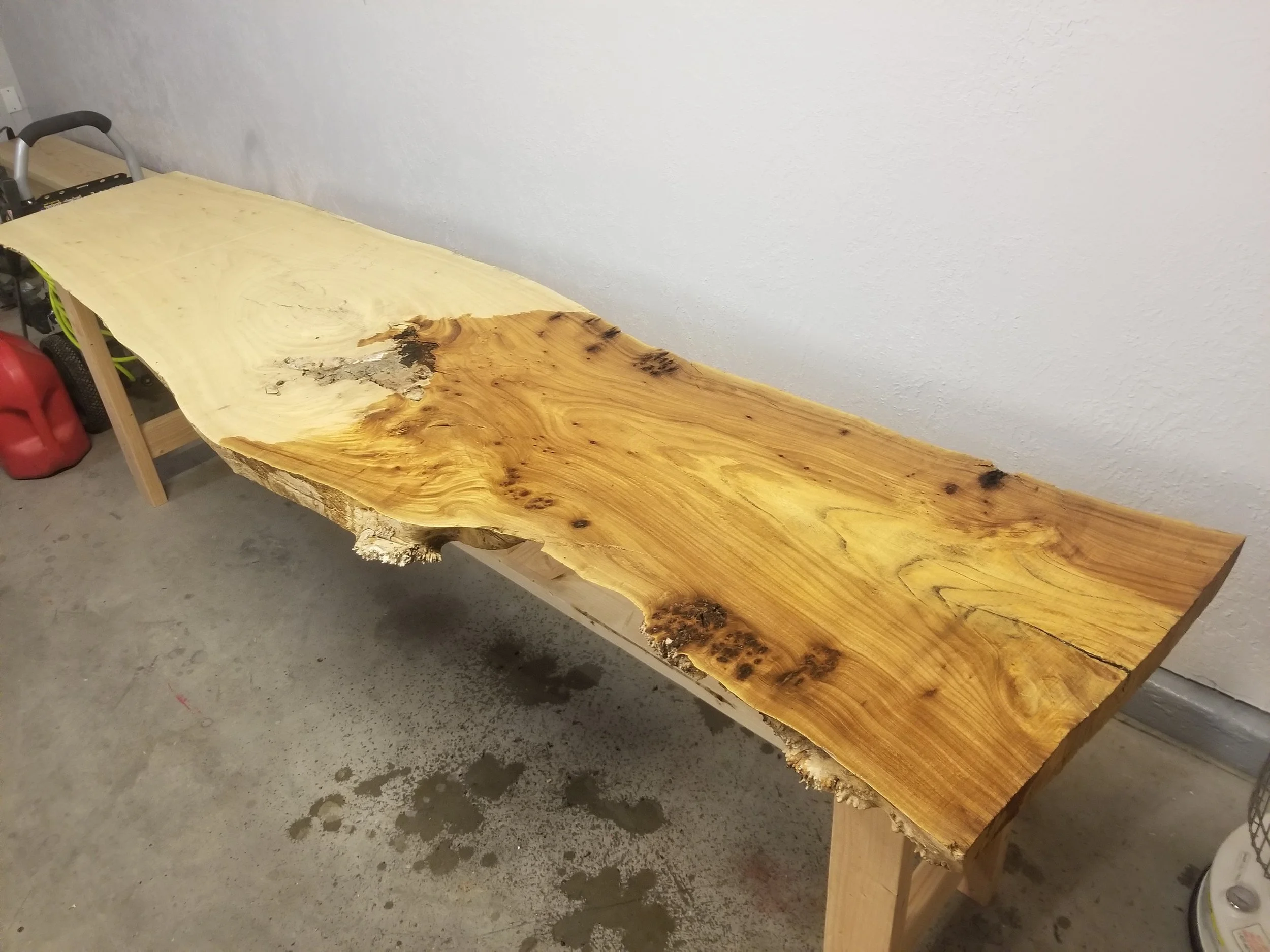 A large, natural wood slab table with a live edge, supported by wooden legs, situated in a room with a gray concrete floor and a plain white wall.
