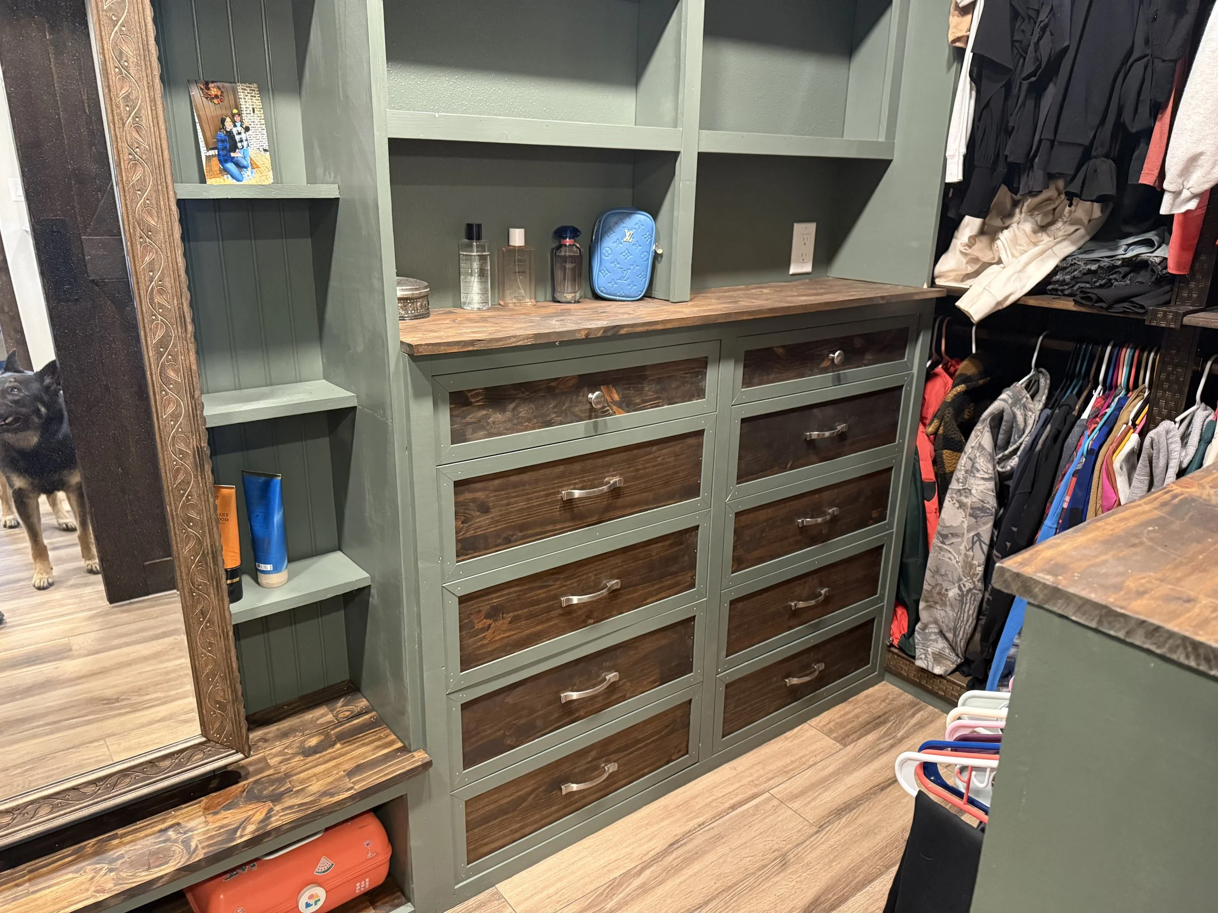 View of an organized closet with green wooden shelves and drawers, some containing bottles of perfume and a blue Louis Vuitton bag. To the right, there are hanging clothes and a rack with colorful hangers. On the left, a partial view of a mirror with