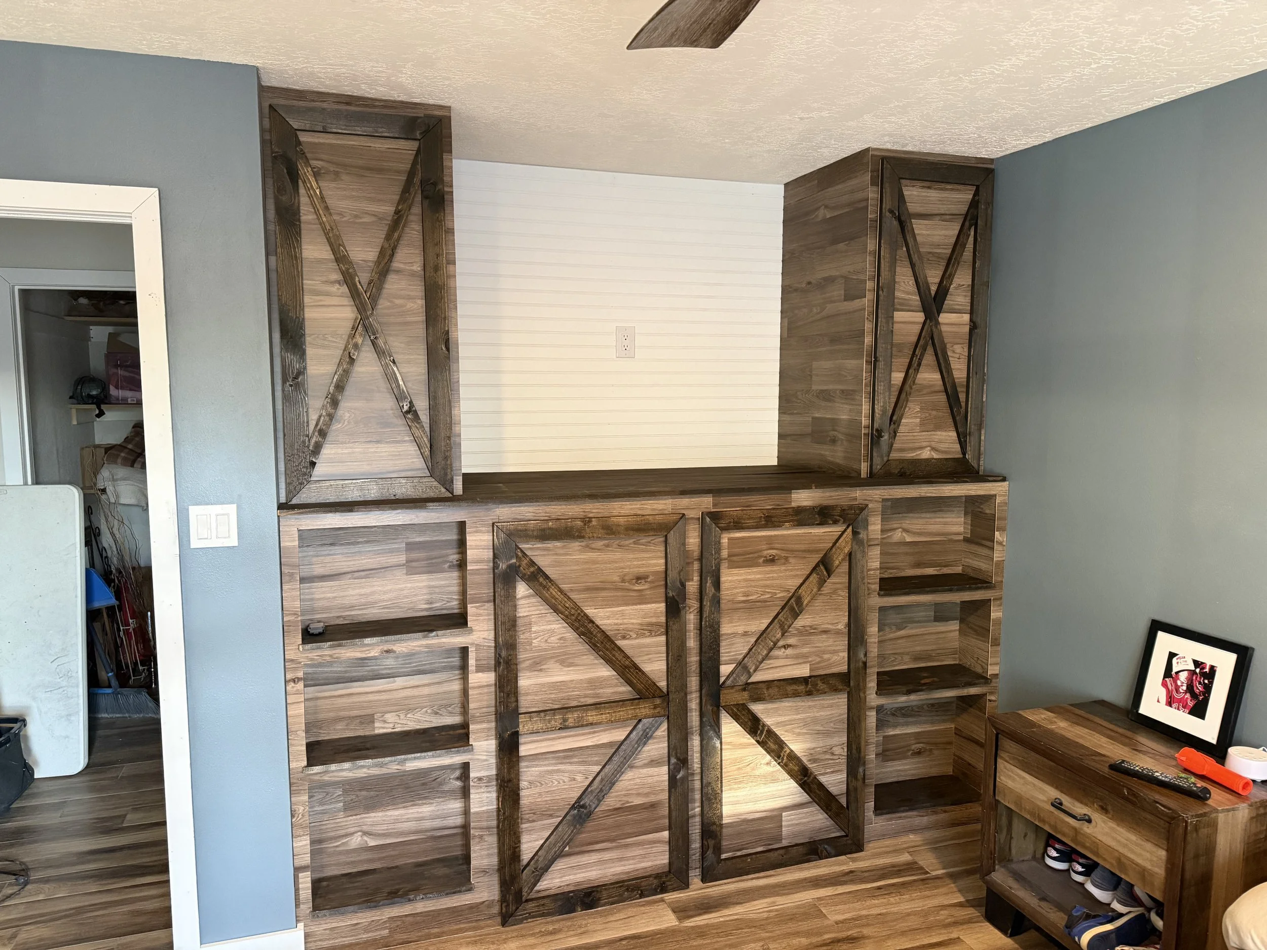 Wooden built in closet, next to a blue wall and a white-panel wall, with a framed photo, a remote, and shoes on a wooden shoe rack.