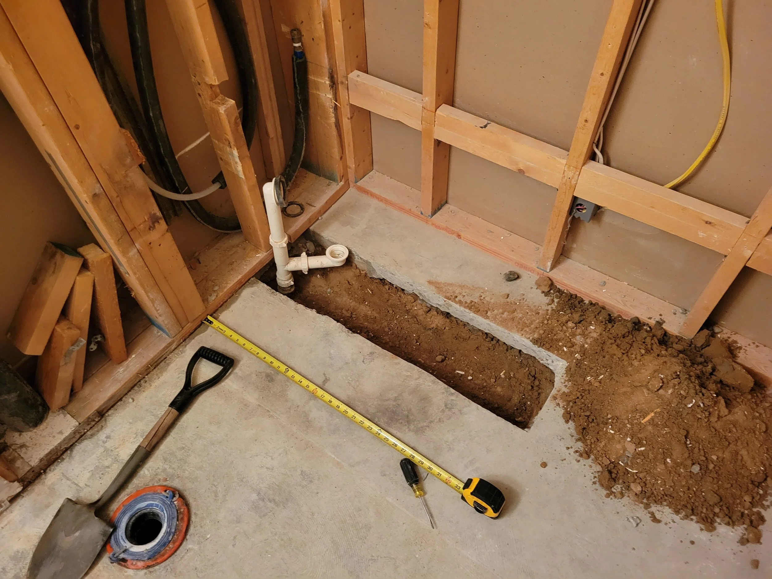 View of a home construction or renovation site showing exposed wooden framing, plumbing pipes, and construction tools on the concrete floor, including a measuring tape, screwdriver, and a roll of tape.
