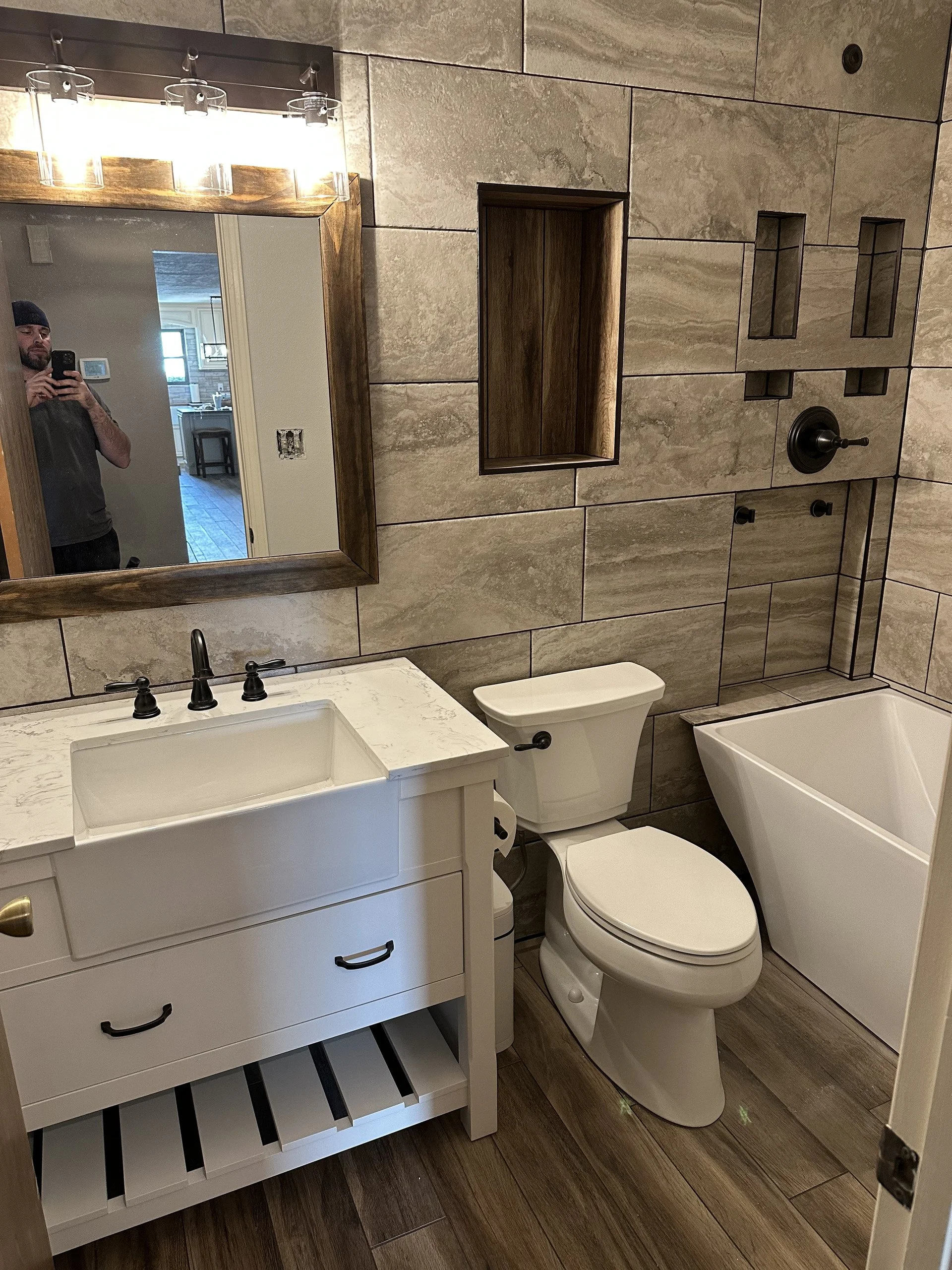 A bathroom with a white vanity with a marble top, a black faucet, a round mirror, a wooden framed medicine cabinet, a modern black toilet, and a bathtub. The wall has large beige tiles with black grout, and the floor has wood-like tiles.