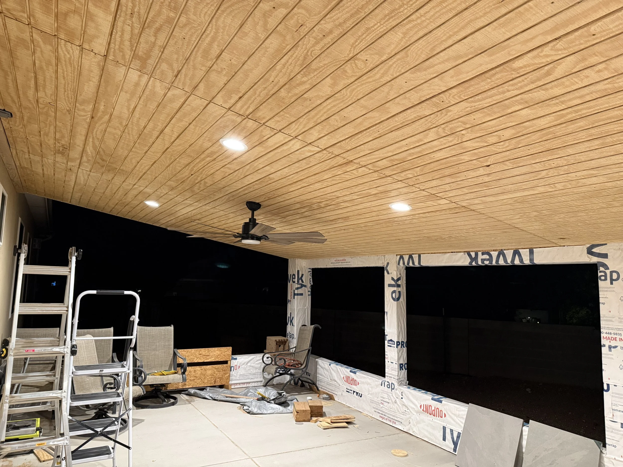 This image shows a covered outdoor area under construction, with a wooden ceiling and integrated lights. Construction materials, tools, and furniture are scattered on the ground, and part of a wall with insulation wrap is visible.