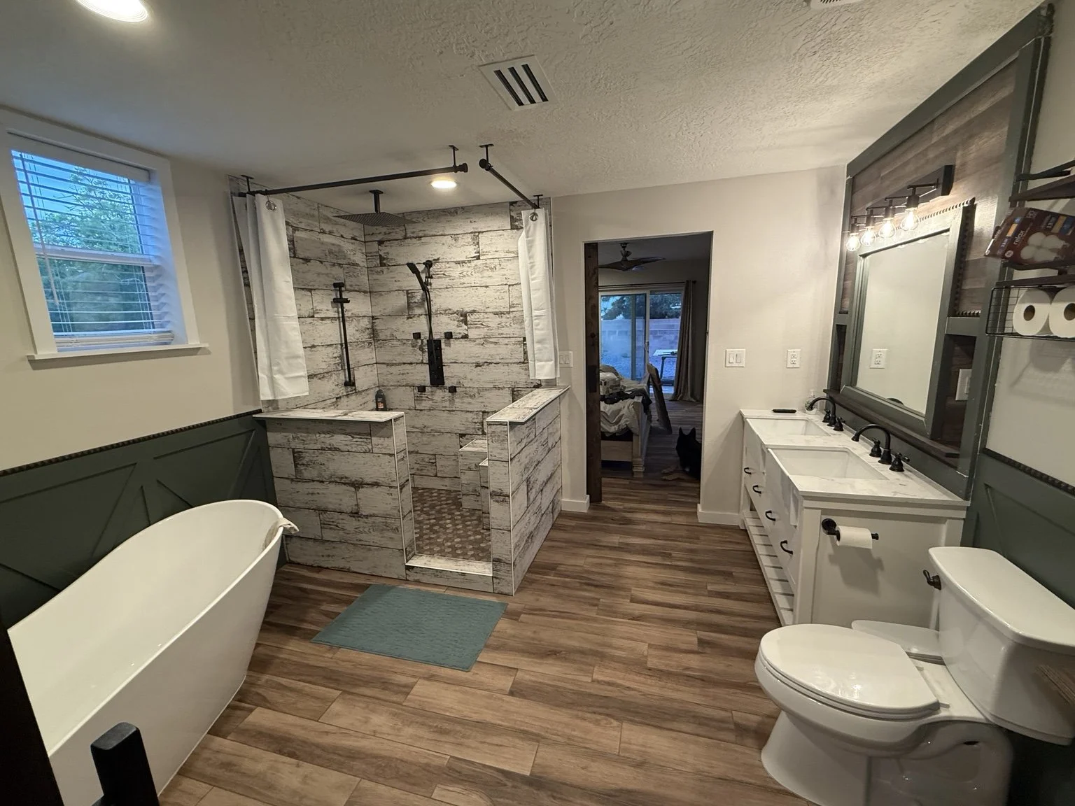 Bathroom with a vintage clawfoot bathtub, a walk-in shower with white and gray wood tile walls, a double vanity with a large mirror, a toilet, wood grain flooring, and a window with blinds.