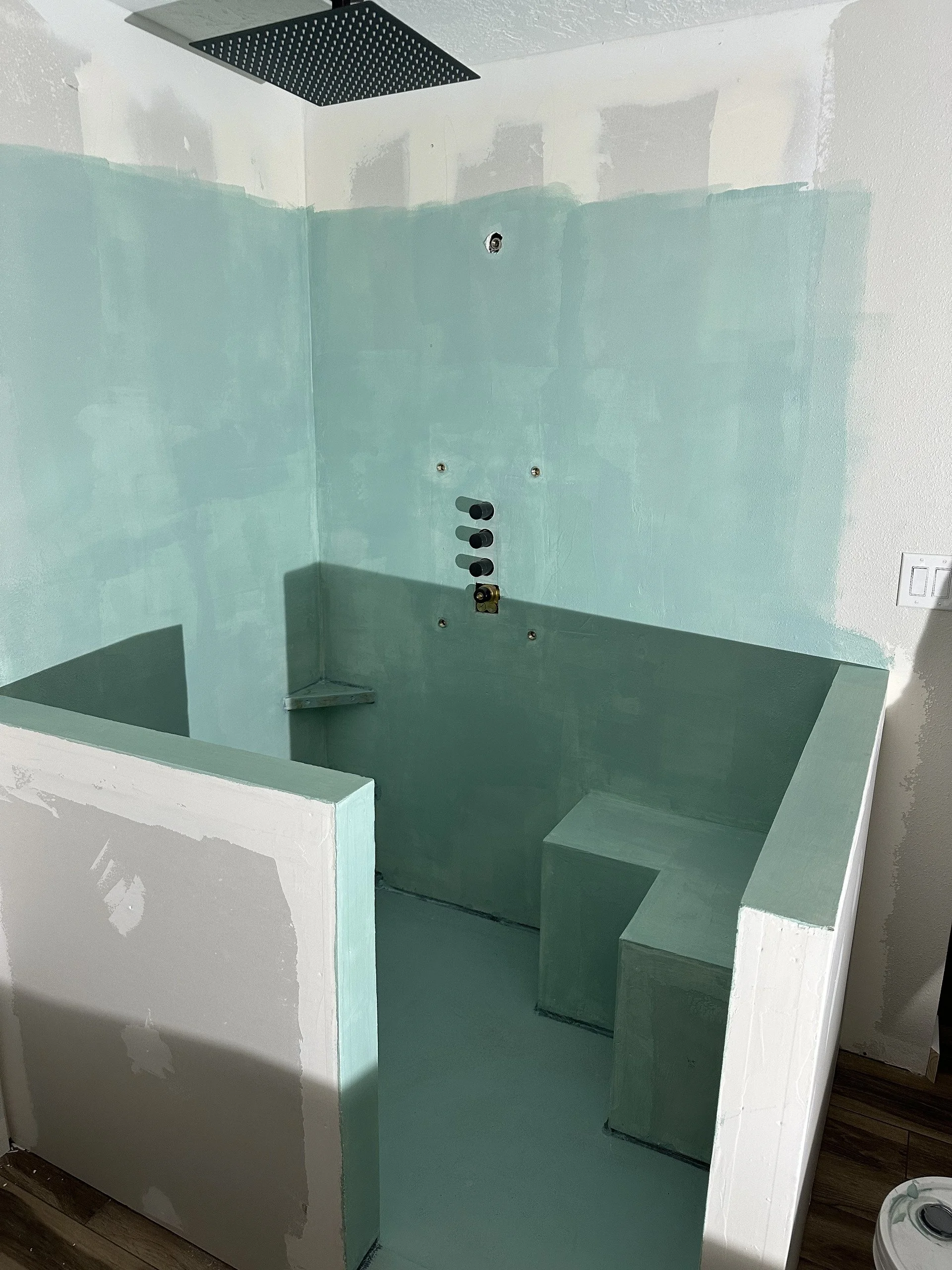 Construction shower with green waterproof walls, black showerhead, and steps, in an unfinished room with drywall and a light switch.