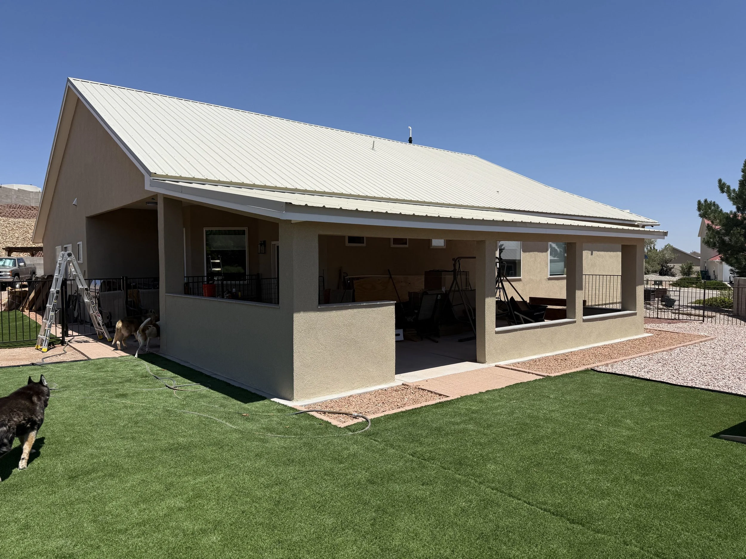 A backyard with a house featuring a beige exterior and a white metal roof. The yard has green grass and a seating area under a covered patio. There are dogs and a ladder visible in the yard, and neighboring houses are in the background under a clear 