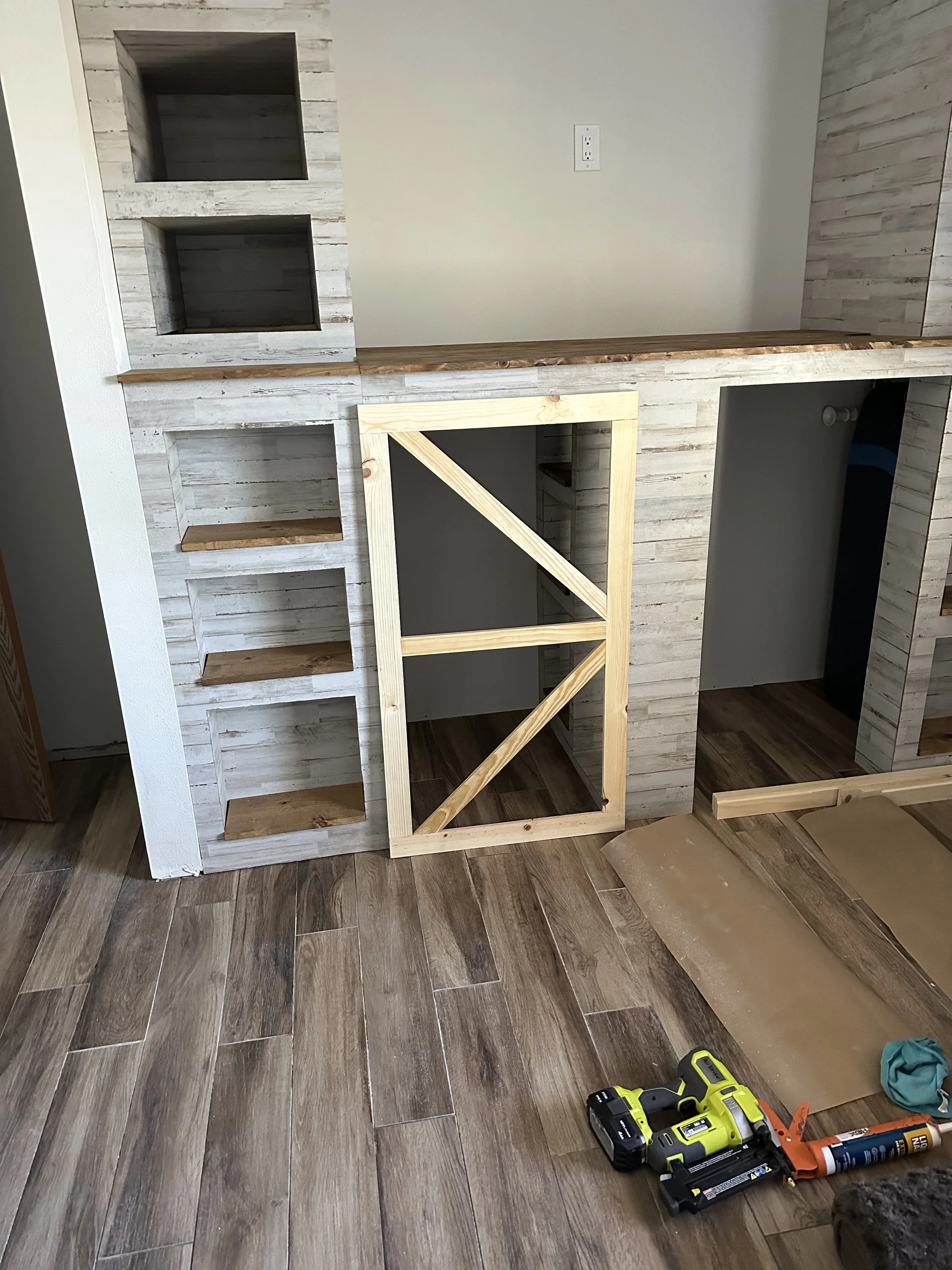 Interior of a room under construction with wooden shelves and a frame, power tools, wood pieces, and a tube of adhesive on a wood floor.