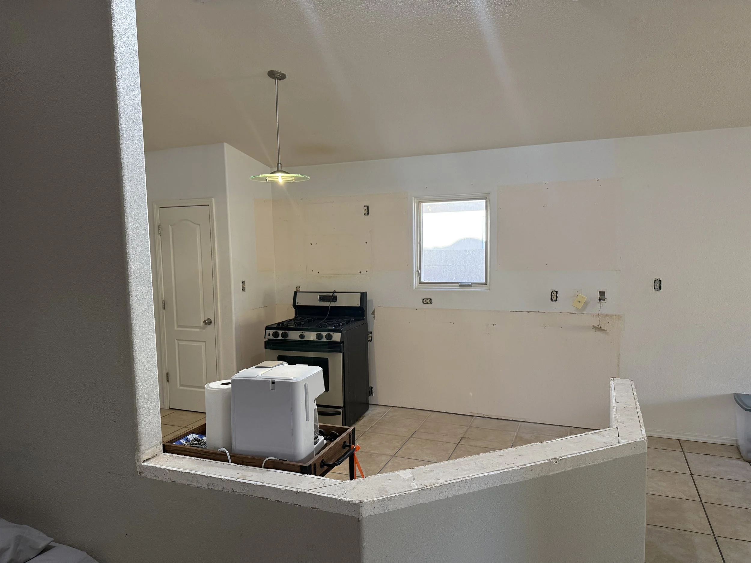 Partially renovated kitchen with white walls, beige tile flooring, and a window letting in natural light. A black stove is placed against the back wall with some electrical outlets above it. The kitchen counter is under construction, with a white app