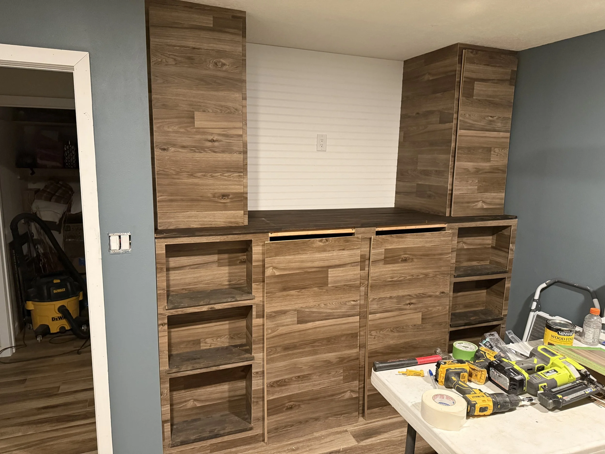 A partially assembled wooden wall unit with multiple open shelves is in a room. To the right, a white table holds various power tools, paint supplies, and tape. The wall behind the unit is painted light blue and has an electrical outlet. A doorway on