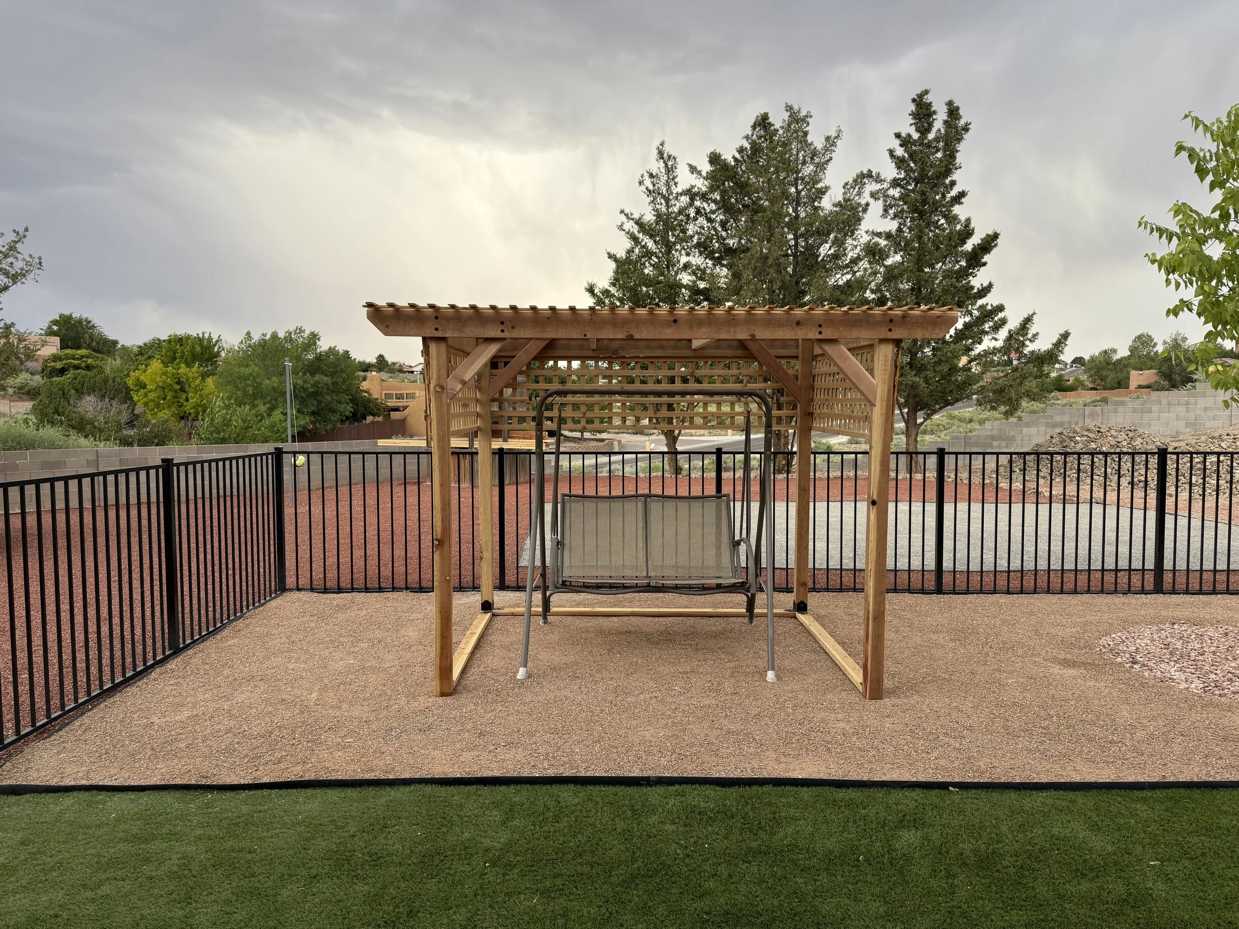 Backyard with a wooden pergola, swing, and fencing, surrounded by trees and greenery under an overcast sky.