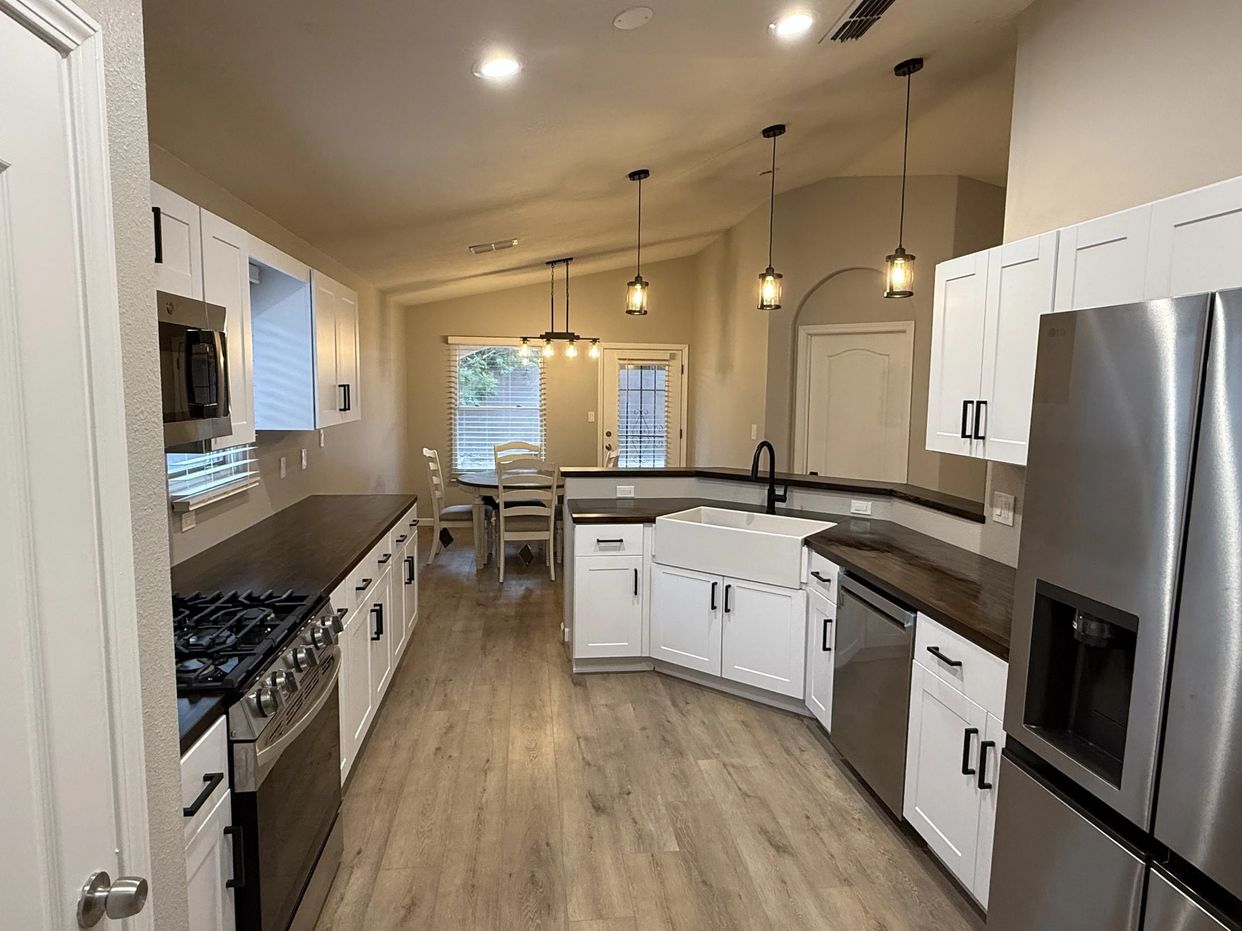 Modern kitchen with white cabinets, dark countertops, stainless steel appliances, and pendant lights hanging from the ceiling. A dining table with chairs is visible through a window with blinds, and there is a door leading outside.