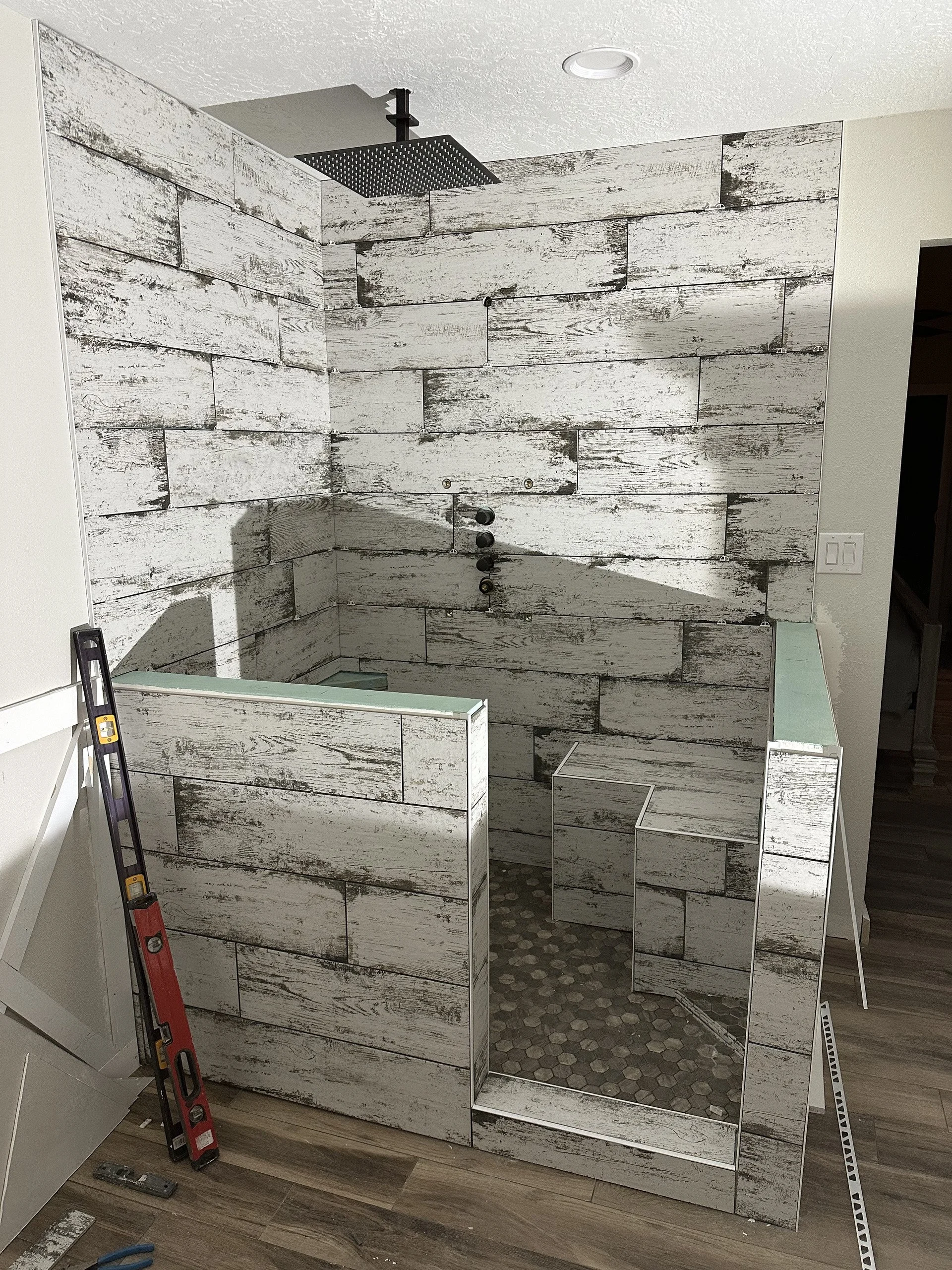 A bathroom under renovation with a partially installed shower wall made of wood-look tiles, a showerhead attached to the ceiling, and construction tools and materials visible on the floor.