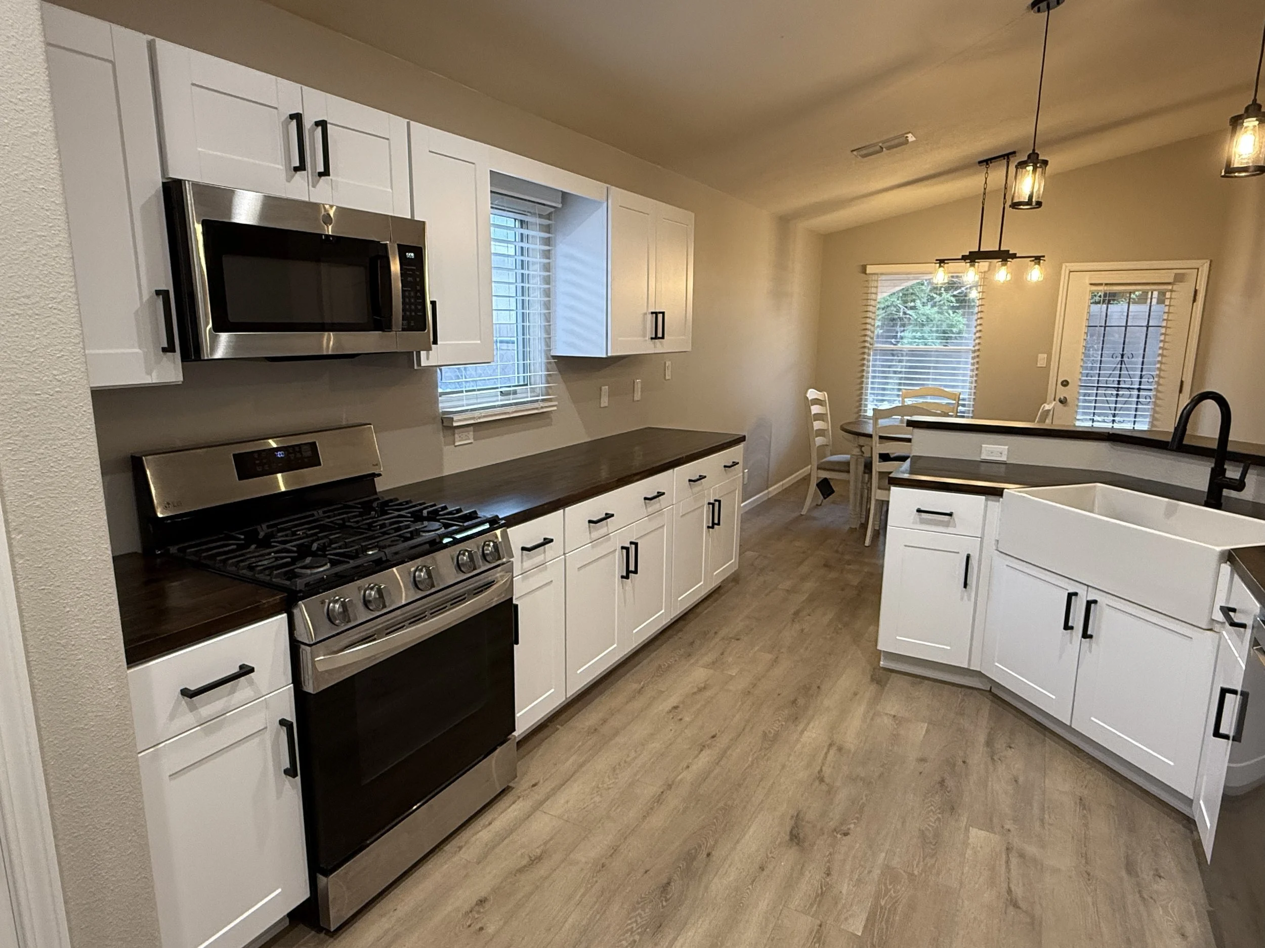 Modern kitchen with white cabinets, stainless steel appliances, and wood flooring. A window and glass door with blinds let in natural light. Overhead hanging light fixtures illuminate the space.