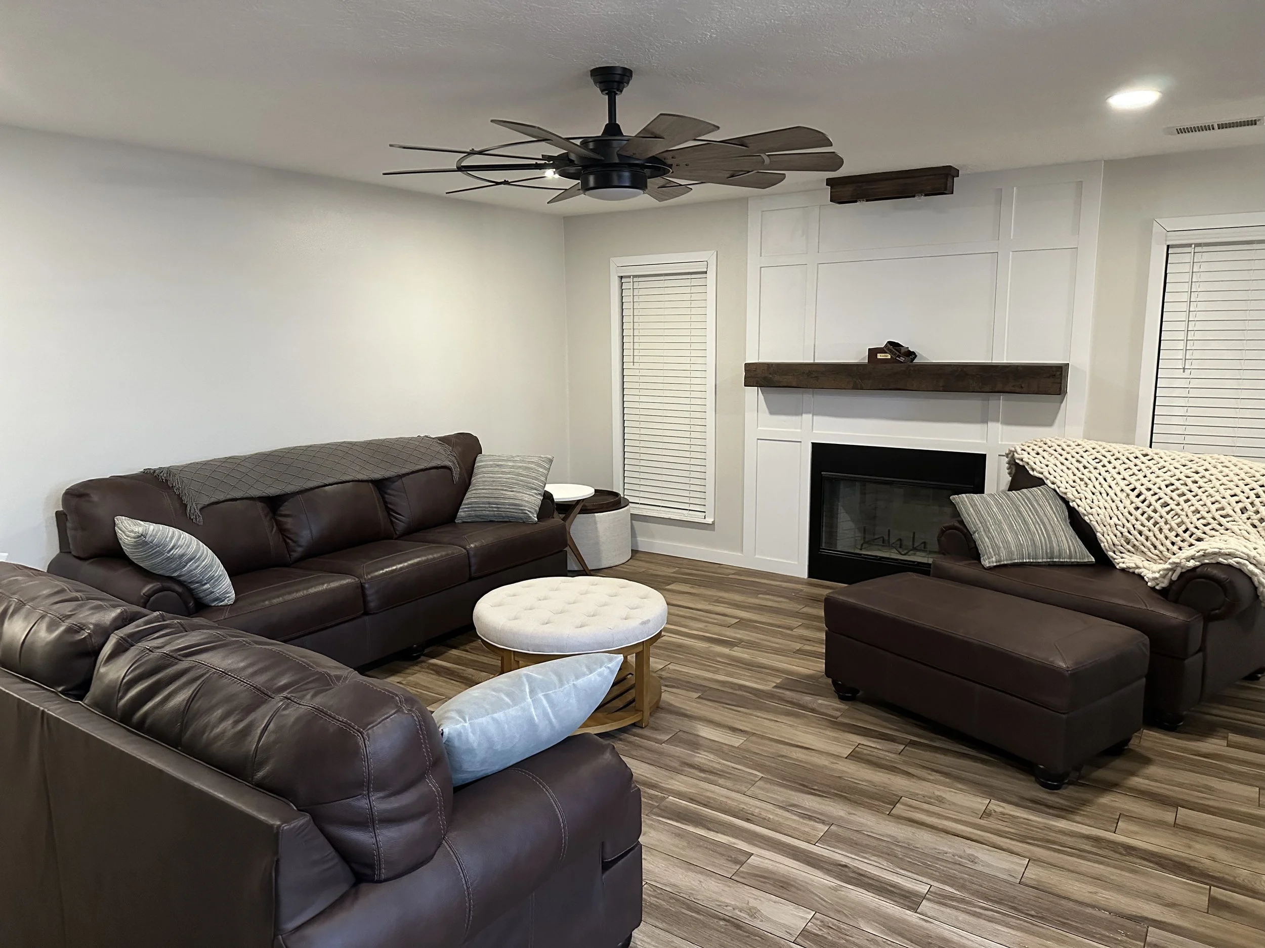 Living room with brown leather sofa, gray pillows, white round ottoman, wooden floors, fireplace, white walls, seagull on brown wood mantel, ceiling fan with dark blades, blinds on windows.