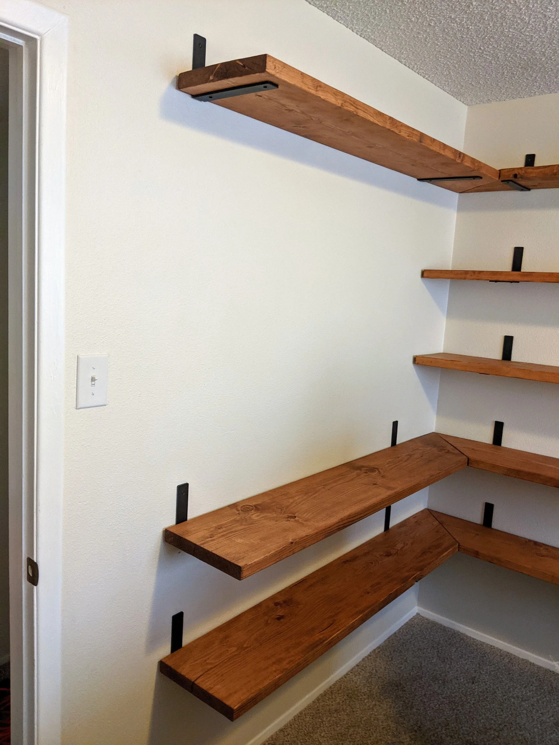 Wooden floating shelves mounted on a white wall in a corner of a room, with black brackets supporting the shelves.