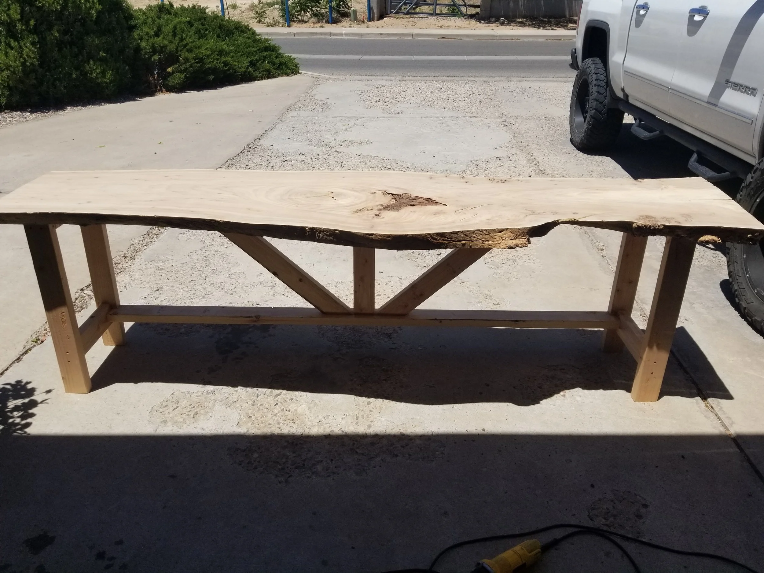 A wooden live edge table with uneven edges on a concrete surface, with a large white truck parked nearby and a sidewalk with bushes and a road in the background.