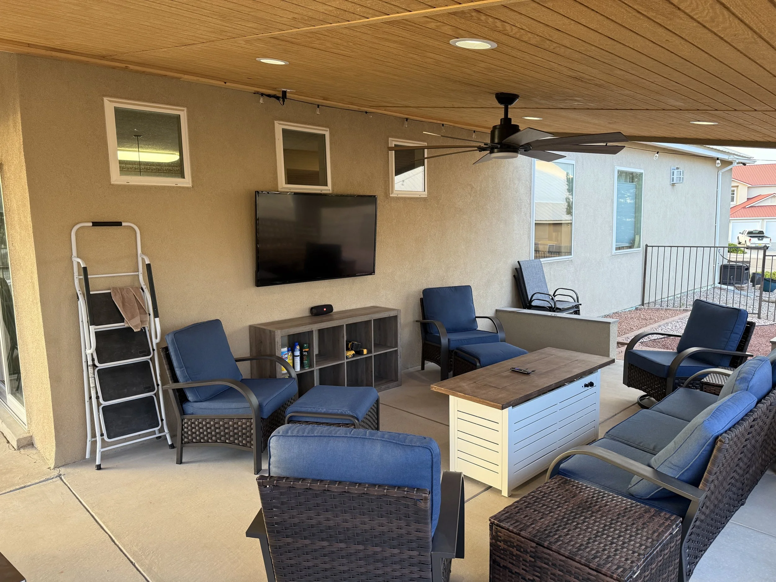 Covered outdoor patio area with ceiling fan, flat screen TV, chairs, a small table, and a bookshelf, overlooking neighboring houses and parked cars.