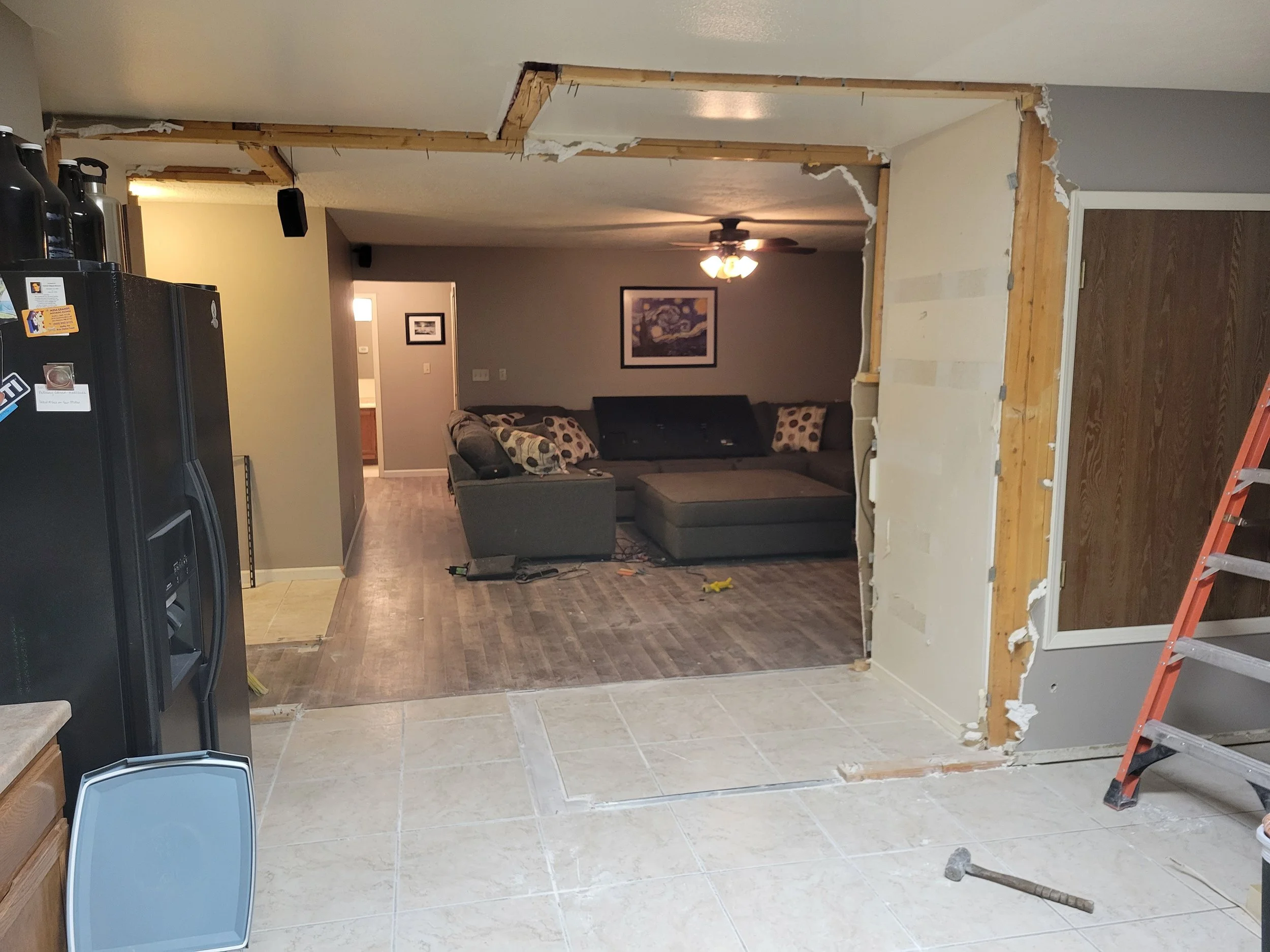 Living room with newly torn down wall, exposing wooden studs and drywall debris, with a gray sectional sofa, framed artwork, ceiling fan, and a partially visible black refrigerator.