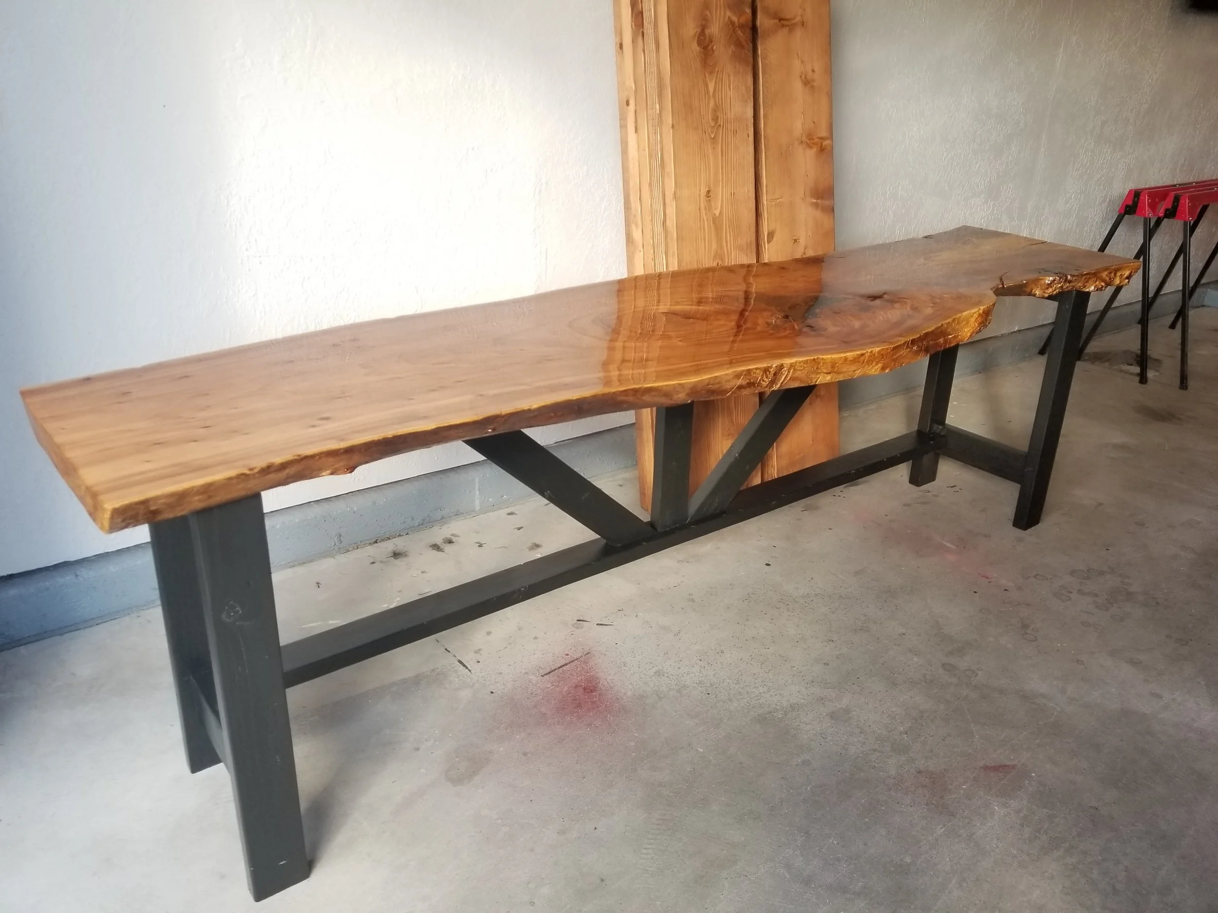 A rustic wooden table with a live edge, supported by black metal legs, positioned against a plain light-colored wall with a small wooden panel behind it. Two black and red stools are nearby on a concrete floor.