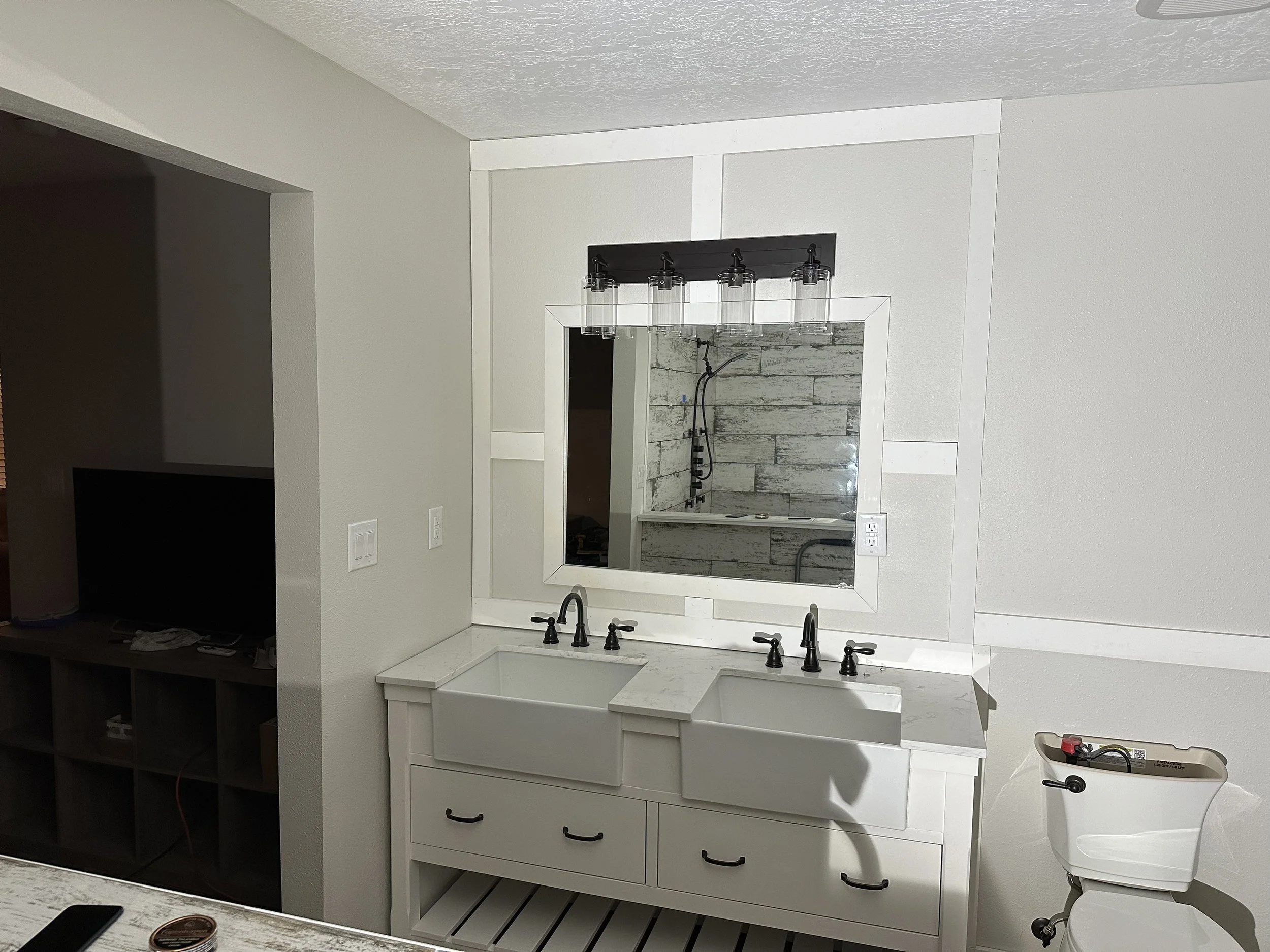 Interior of a bathroom under renovation with a white farmhouse sink, black fixtures, a wall mirror, and a partially tiled wall. A black chandelier hangs above the sink.
