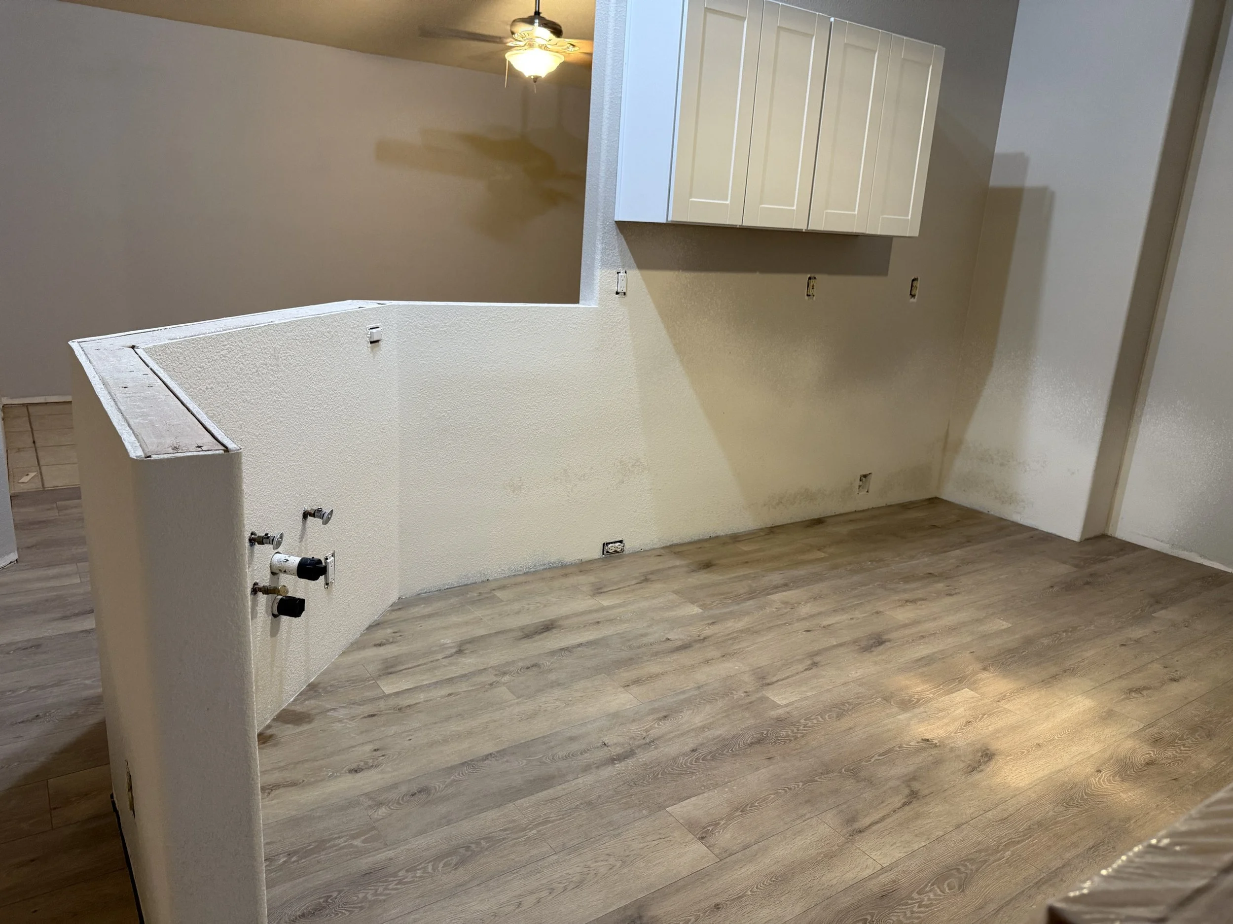 Unfinished kitchen space with exposed pipes and electrical wires, beige walls, white cabinets, hardwood flooring, and a ceiling fan with light.