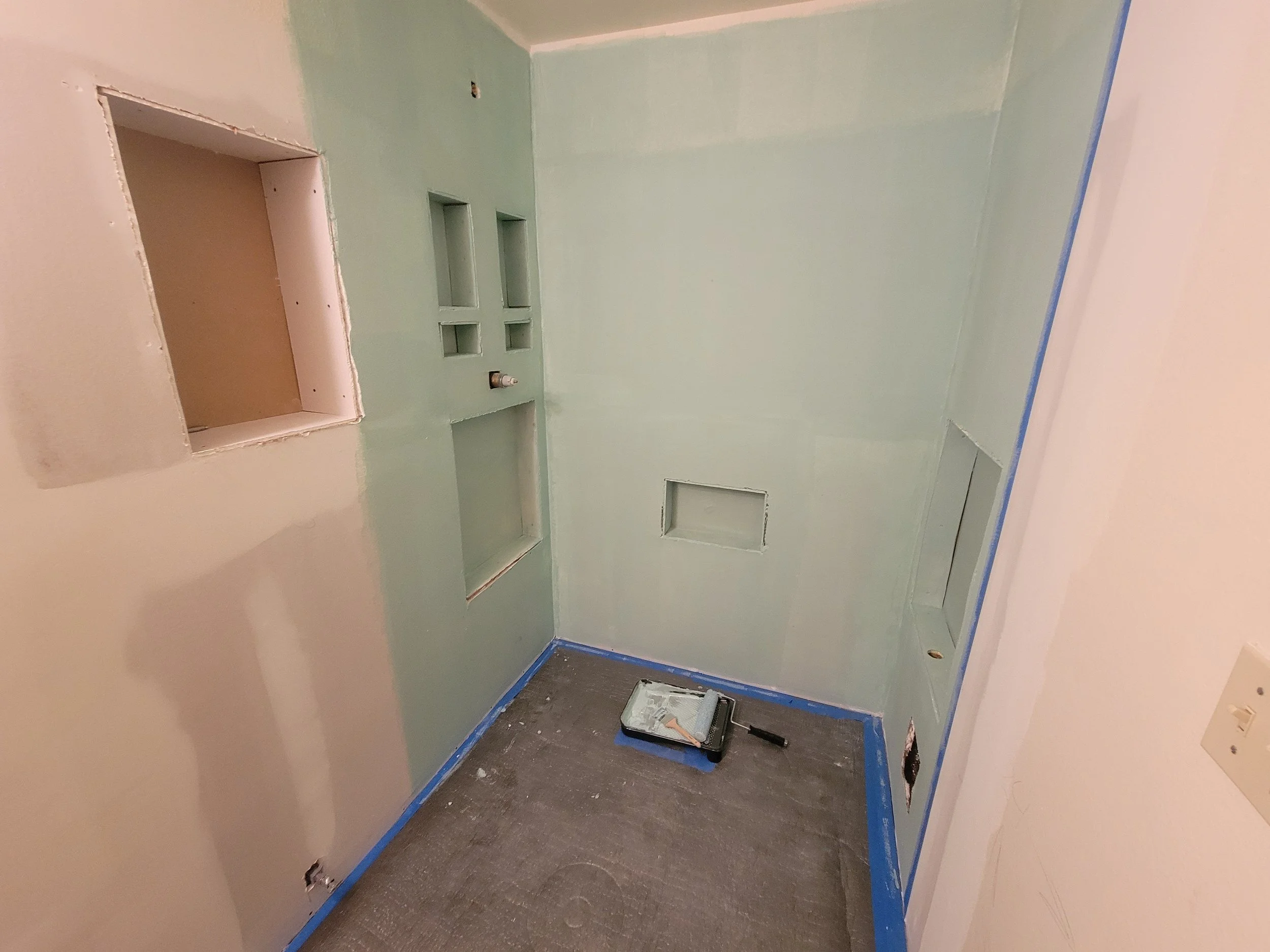 Partially completed bathroom or kitchen wall with paneling and openings for fixtures; walls are being painted light green, with painter's tape along the edges and a small paint tray with paint roller on the dark brown flooring.