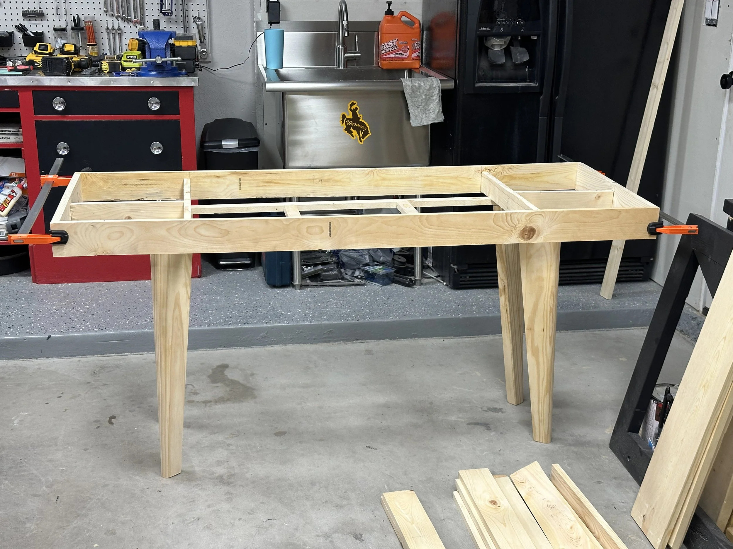 A workshop with a wooden table frame clamped to a workbench, various tools on the workbench, a black recycling bin, a stainless steel sink with a blue cup and orange container, and a black refrigerator. A sticker of Wyoming is on the sink.