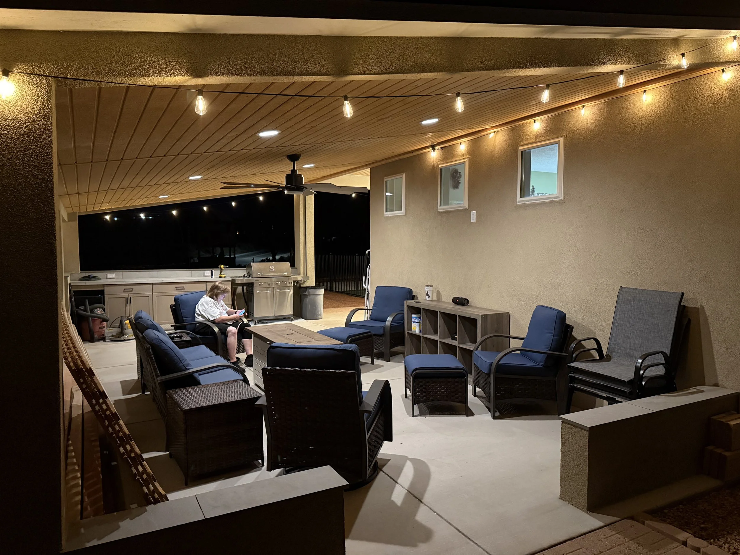Nighttime outdoor patio with string lights, ceiling fan, windows, and seating area with chairs and a woman sitting and using her phone.