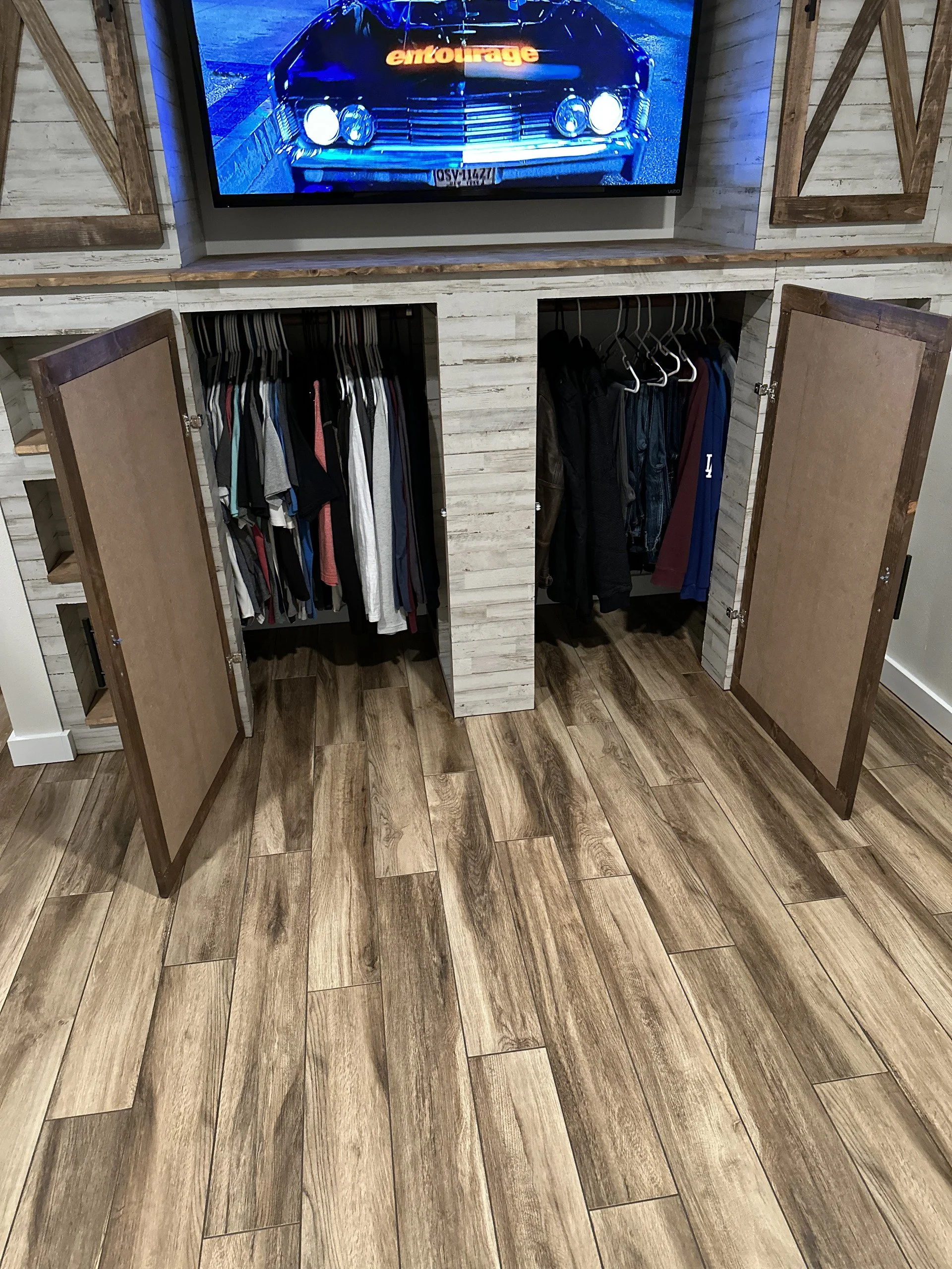 A closet with wood doors open, showing hanging clothes inside, and a flat-screen TV mounted above it.