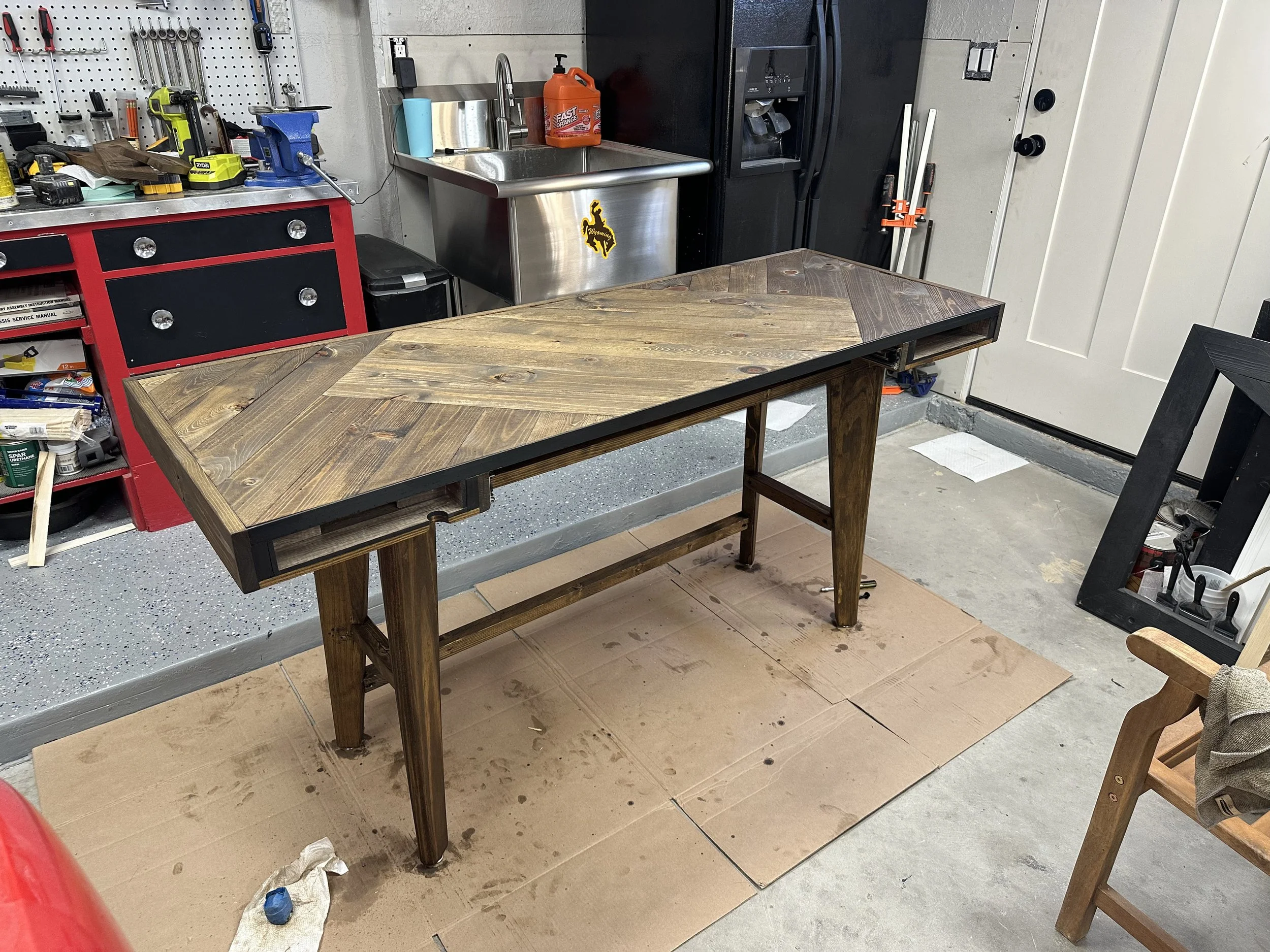 A workshop with a wooden table, a pegboard with tools, a black refrigerator, a stainless steel sink with orange hand soap, and a partially visible door.