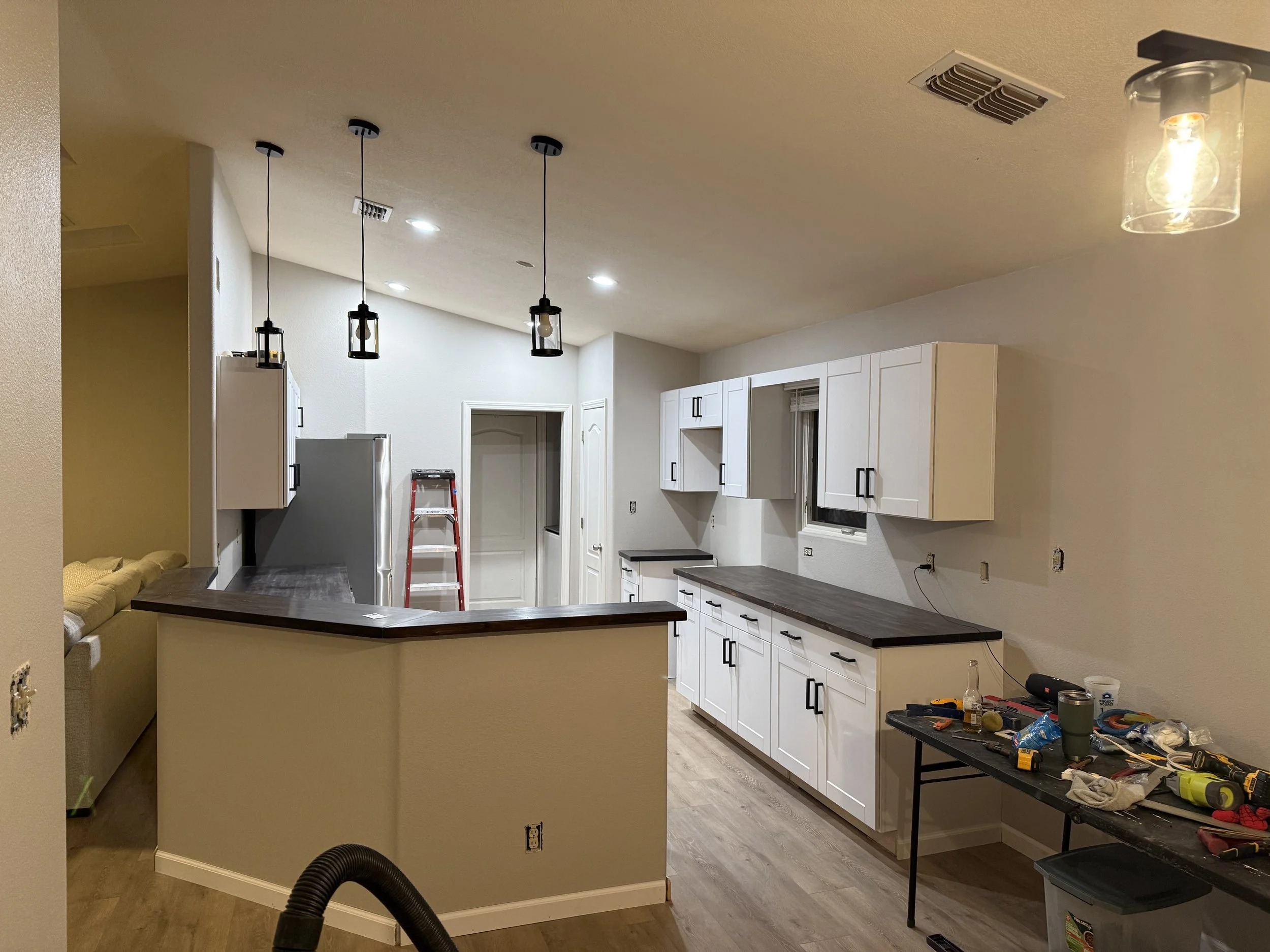 A kitchen under renovation with white cabinets, a countertop, and construction tools on a black worktable. There are three hanging light fixtures and a ladder in the background.