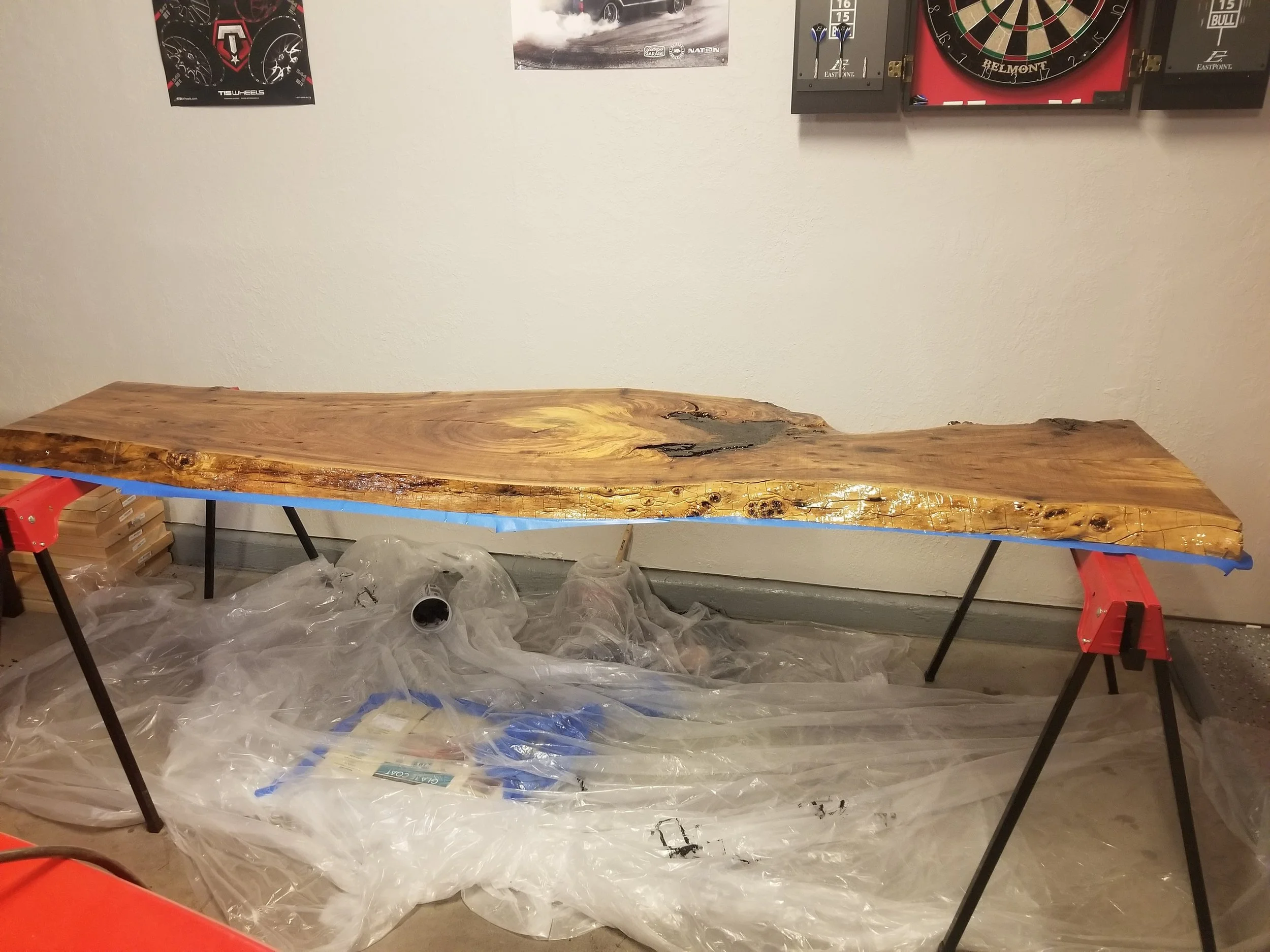 A long, polished wooden slab resting on two orange saw horses in an indoor workshop or garage. The wall behind features posters of car racing and a dartboard. Plastic sheets and packaging are visible underneath and around the workspace.