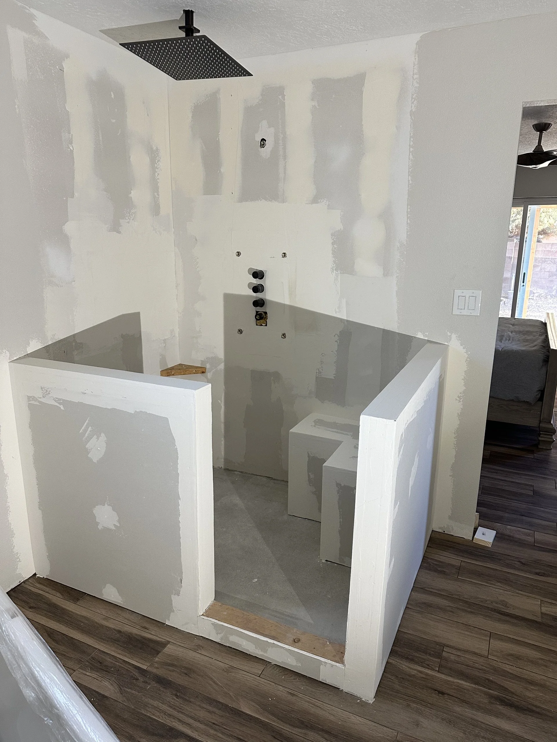 A bathroom in the process of renovation. The shower area has drywall installed, with visible unpainted joint compound and screw holes. The shower has a black overhead rainfall showerhead. The surrounding walls are white, and the flooring is wood-like