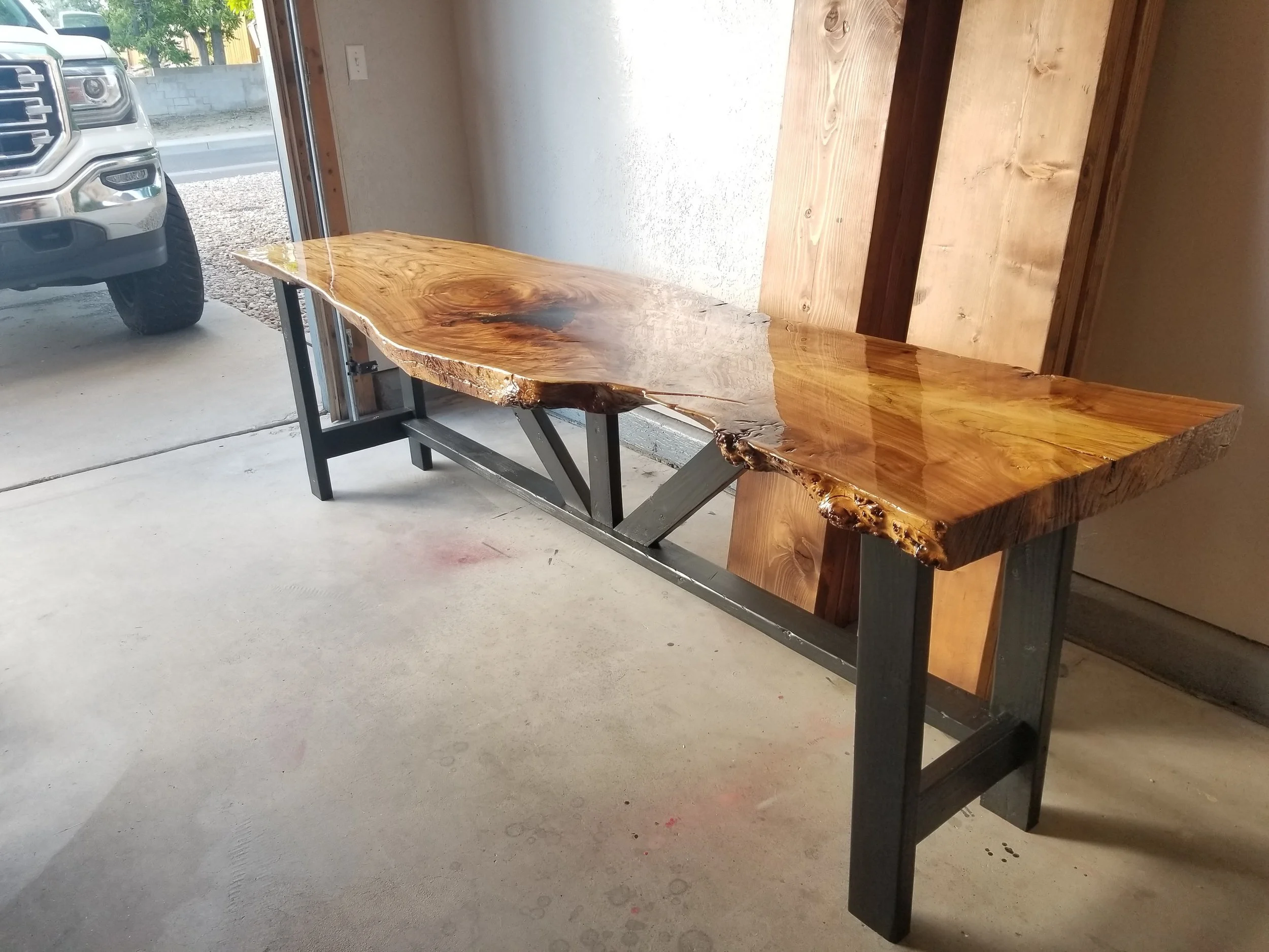 A live edge wooden table with a polished surface and natural wood grain, supported by metal legs, situated near a large glass door showing parked vehicles outside.