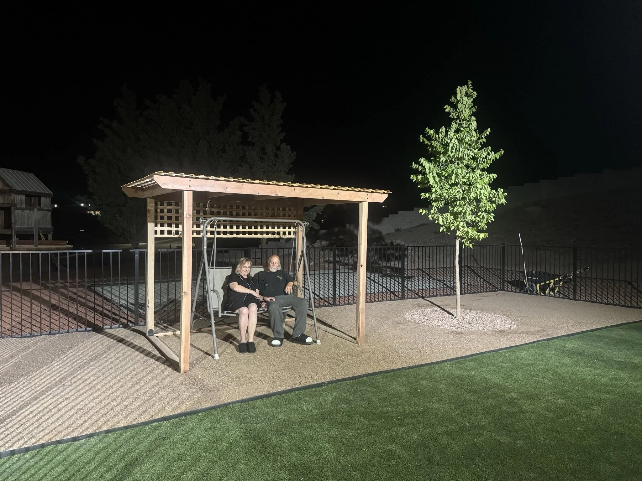 Nighttime scene with a couple sitting on a swing under a wooden gazebo, surrounded by a black metal fence, with trees and a playground in the background.