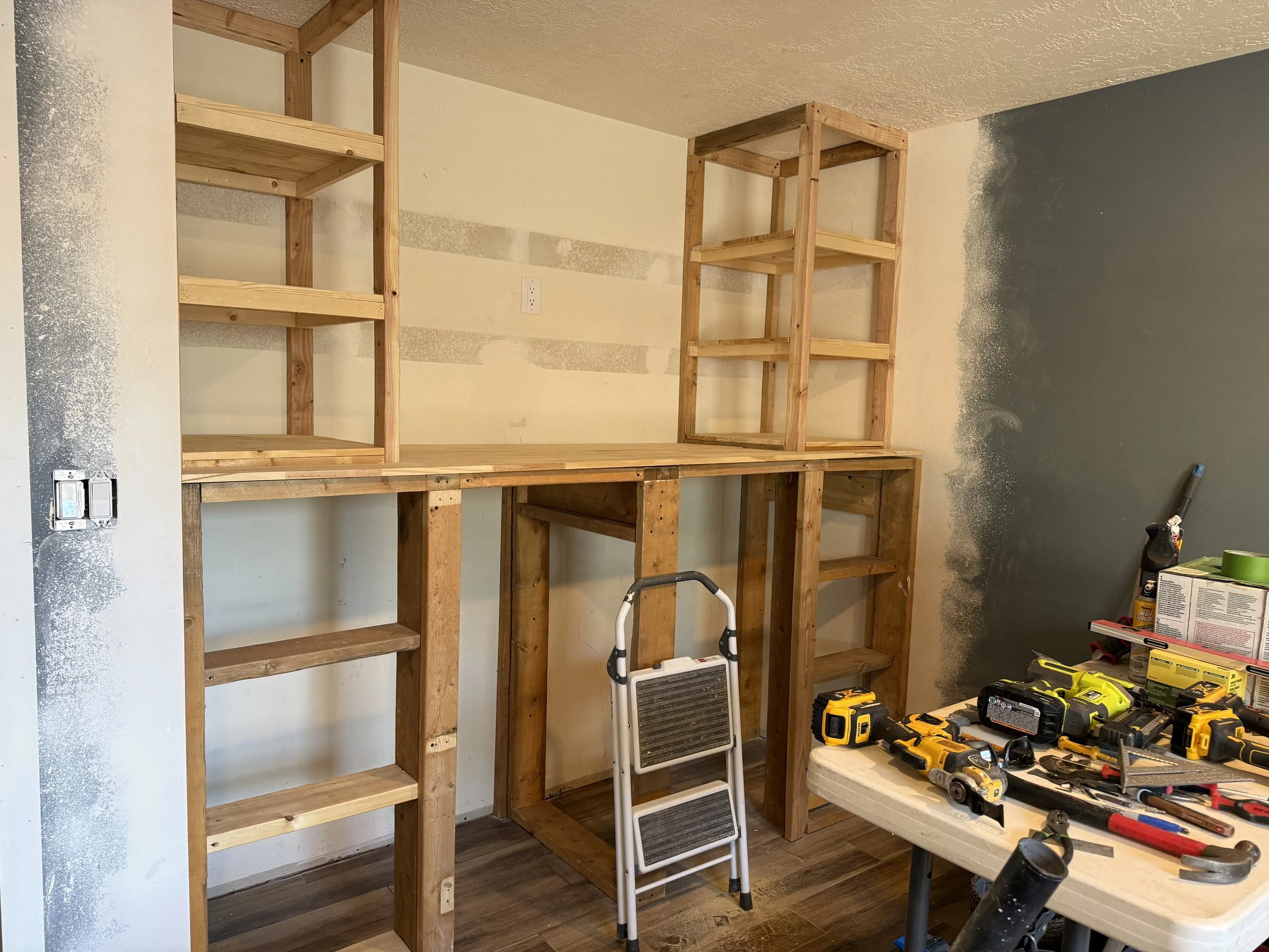 Photo of a closet with wooden framing and a single painted wall on the right. There are tools and supplies on the table in the foreground, including cordless drills, a level, and paint cans. A step ladder is positioned in front of the framing.