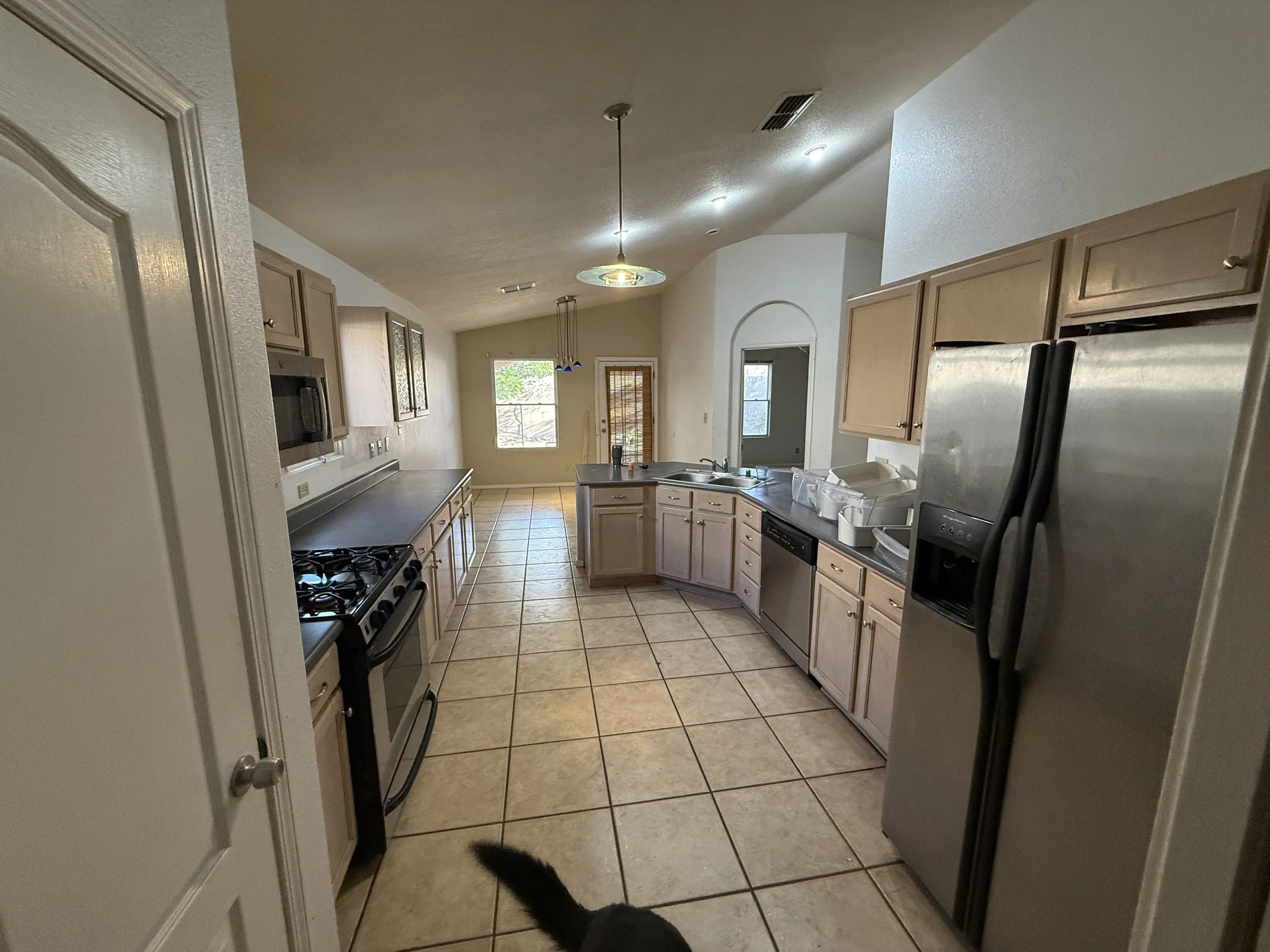 Kitchen with beige cabinets, stainless steel appliances, tile floor, and a ceiling light fixture. A black cat's tail is visible at the bottom of the image.