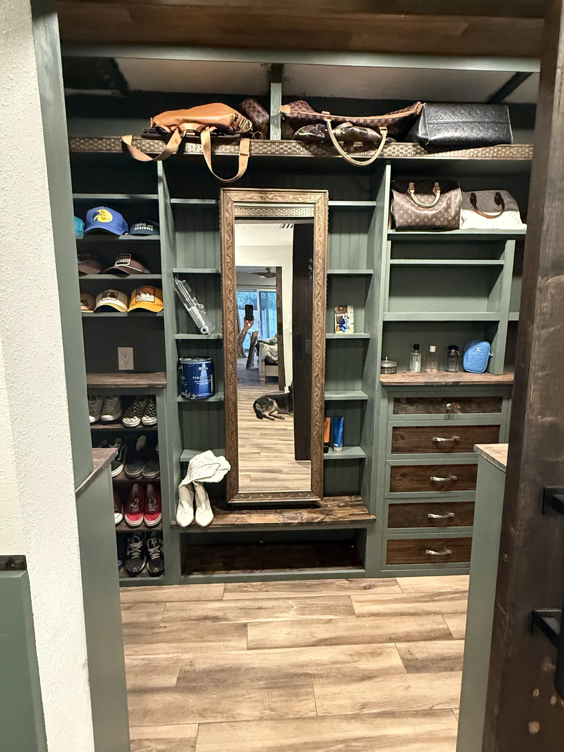 The image shows a greenbuilt-in closet with multiple shelves, drawers, and a full-length mirror. Several handbags are placed on the top shelf, and a pair of shoes and other miscellaneous items are on the lower shelves. In the reflection of the mirror