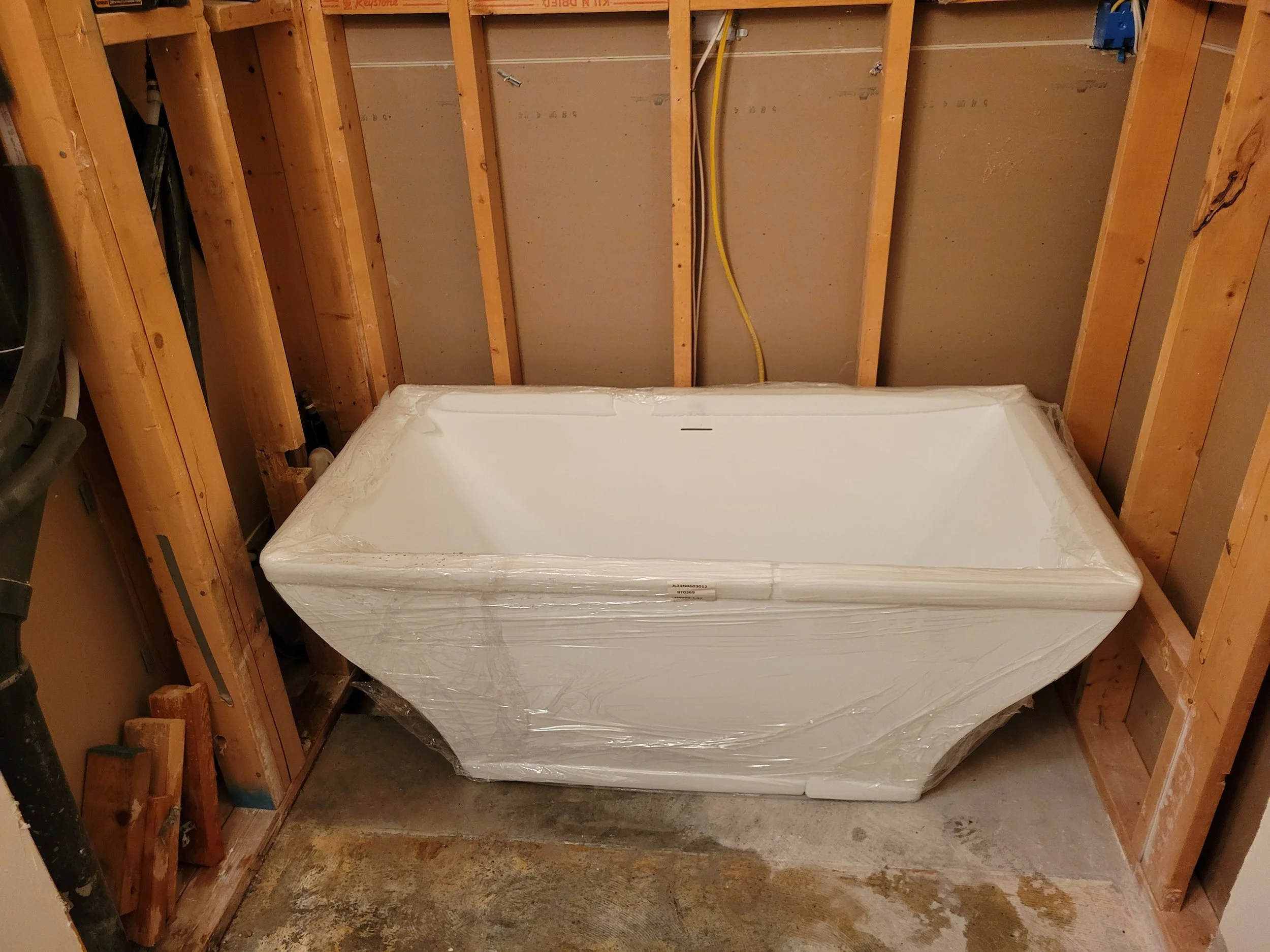 Construction site with exposed wooden framing and a sealed white appliance, possibly a dishwasher or refrigerator, wrapped in plastic.