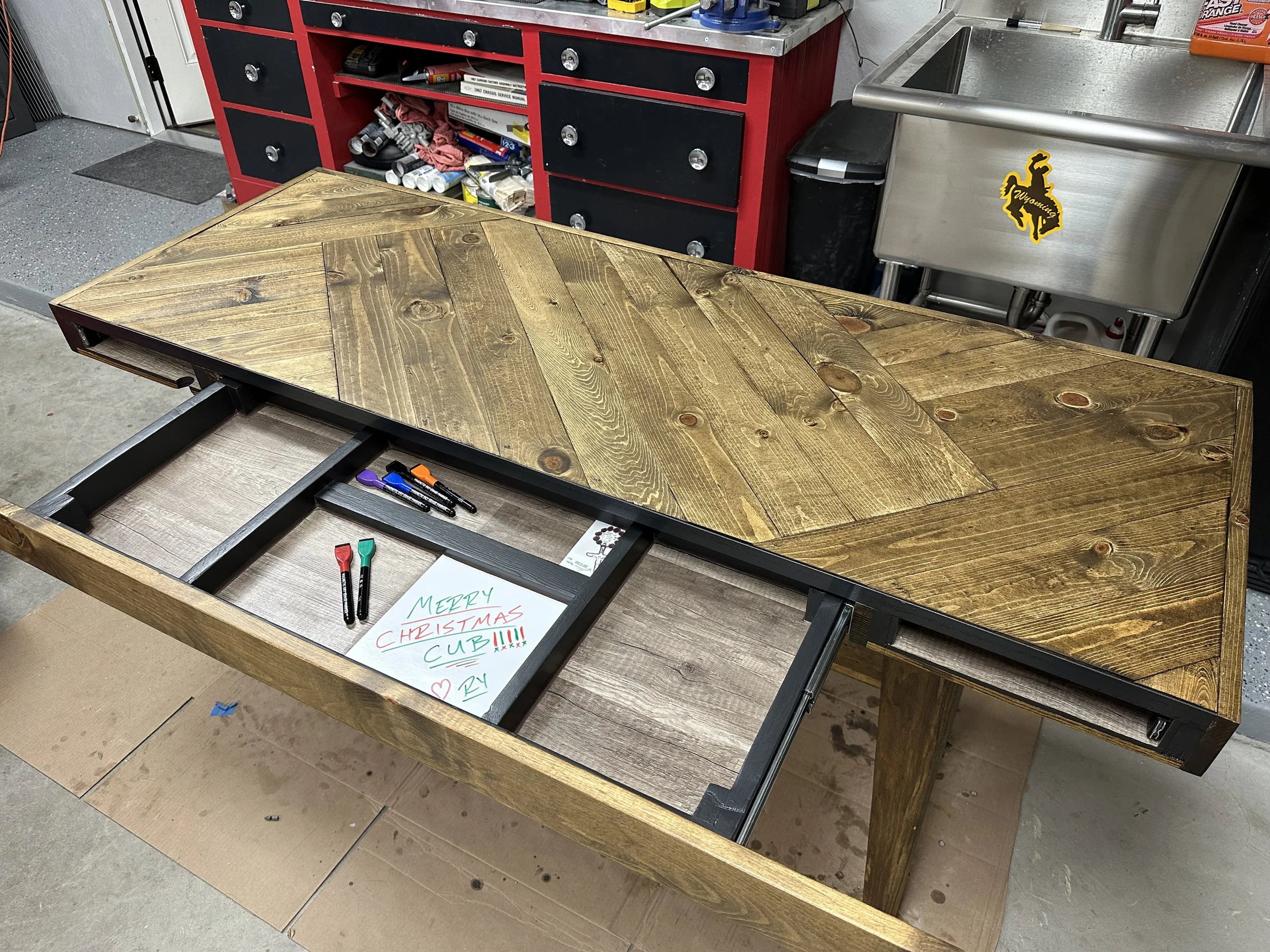 A wooden table with a black frame in a workshop, with colored markers and a handwritten sign that says, 'MERRY CHRISTMAS CUB!!!' The background includes a red and black tool chest, a stainless steel sink with a Wyoming sticker, and various tools and 