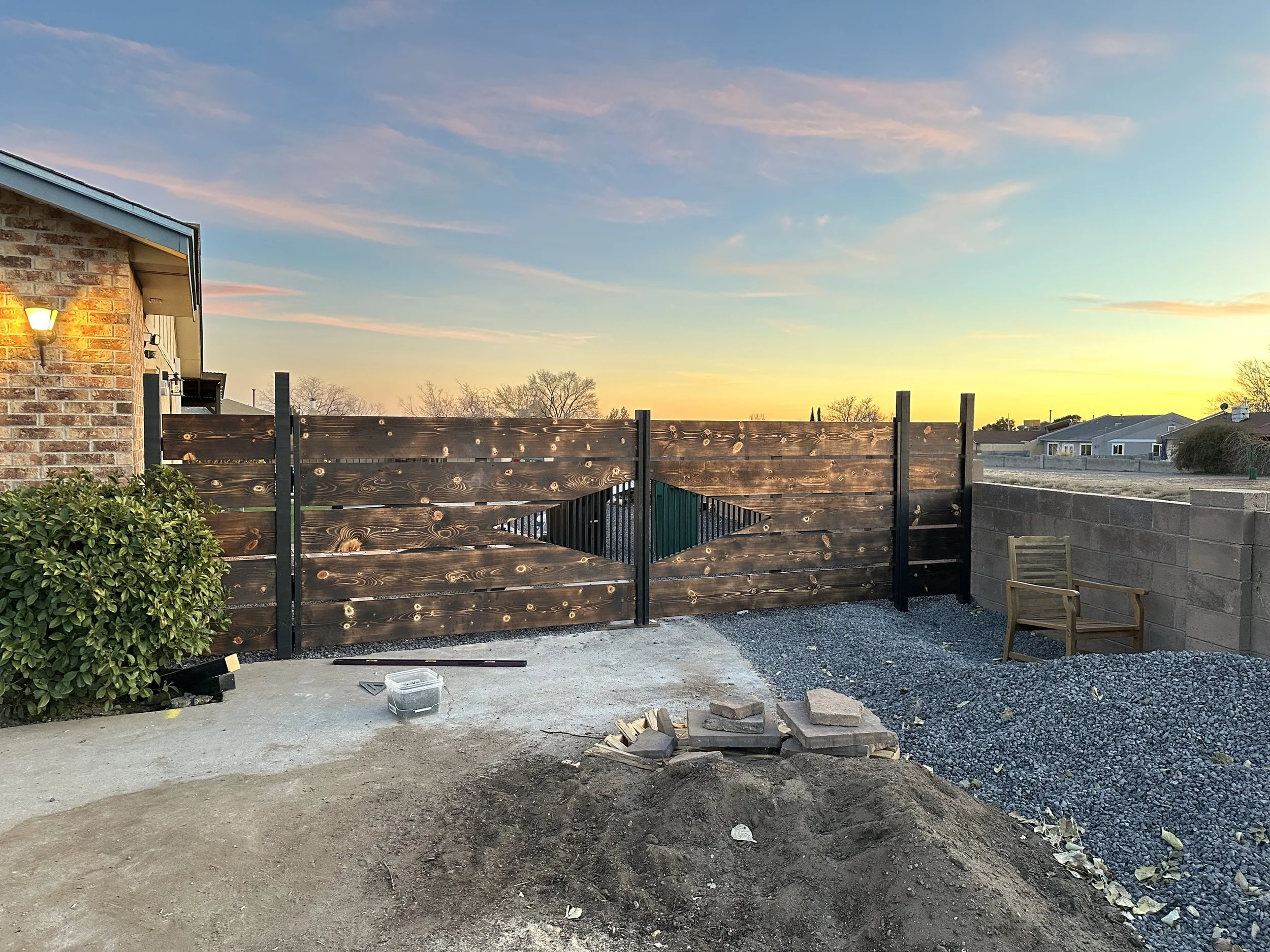 View of a backyard area during sunset, showing a wooden gate, outdoor chairs, a brick house wall with a light, and a yard under construction with gravel and paving stones.