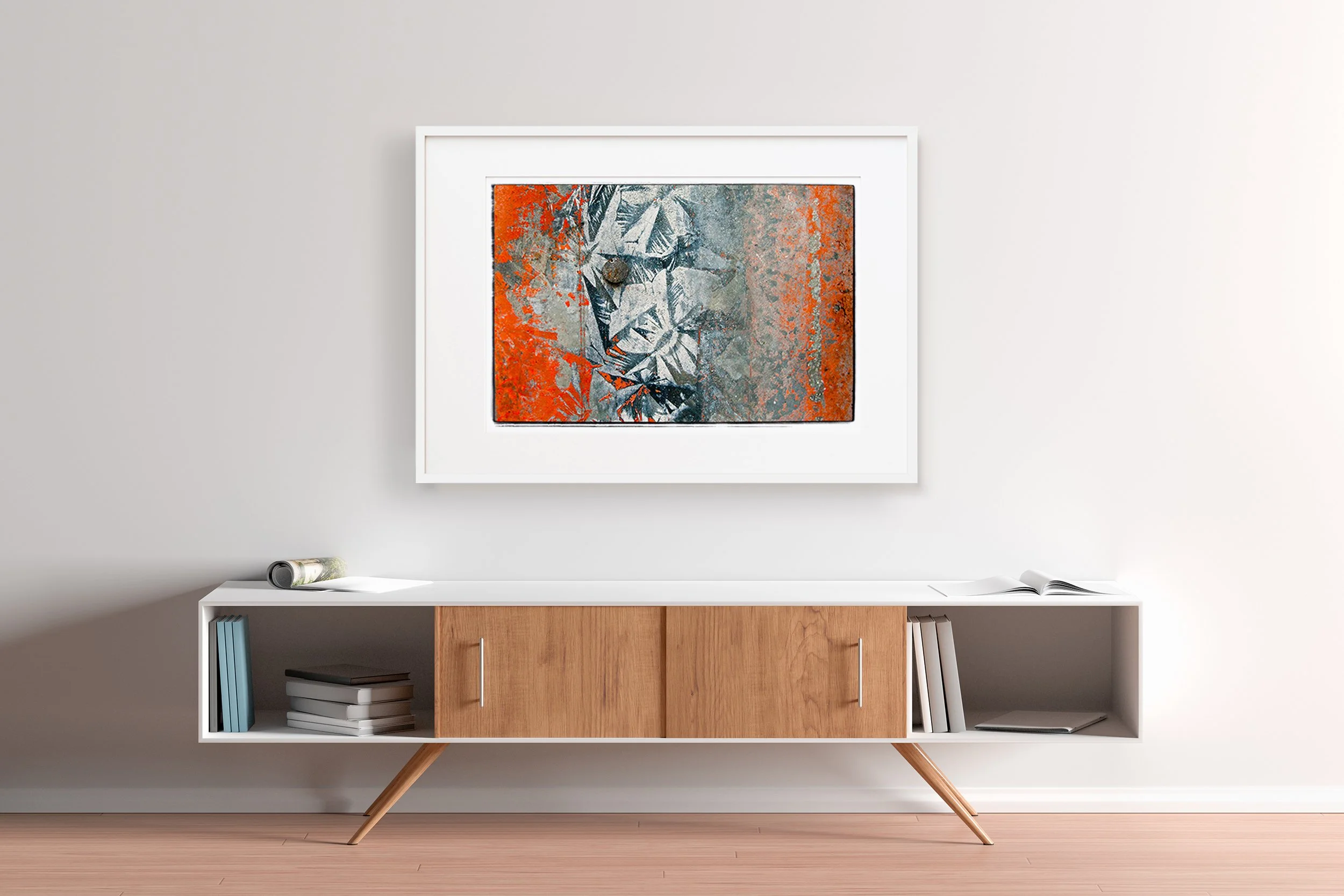 Modern wooden sideboard with white top and angled legs against a plain white wall, decorated with a colorful abstract art piece framed in white hung above, and books and magazines placed on and inside the sideboard.