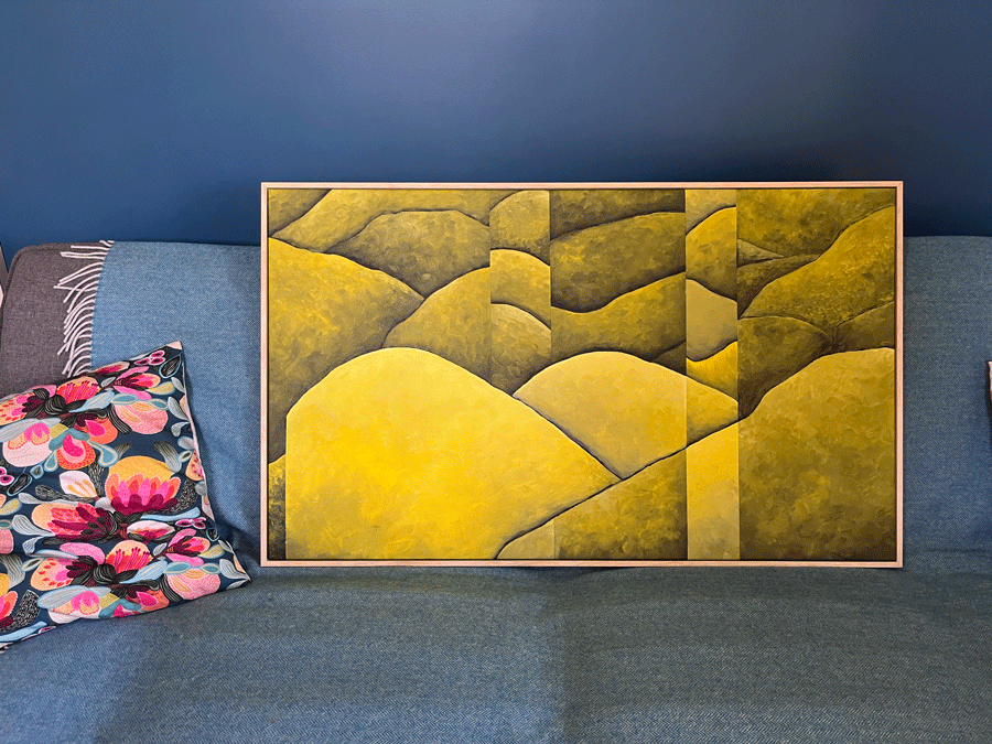A framed painting of green hills on a blue wall, resting on a blue sofa with a floral throw pillow nearby.