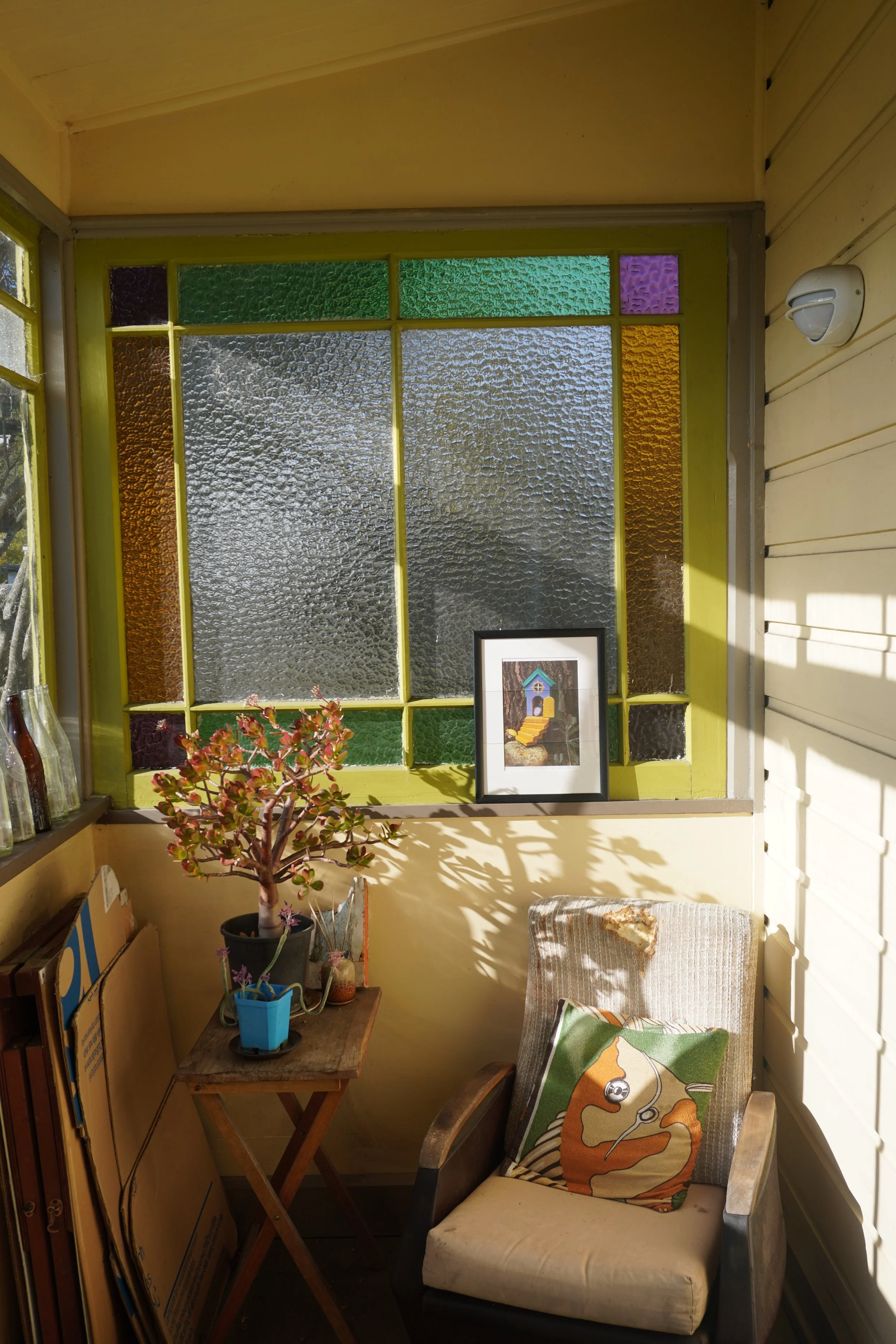 A cozy corner with a wooden armchair with a bear pillow, a small folding wooden table with potted plants, and a stained glass window with colorful textured panes, surrounded by yellow walls.