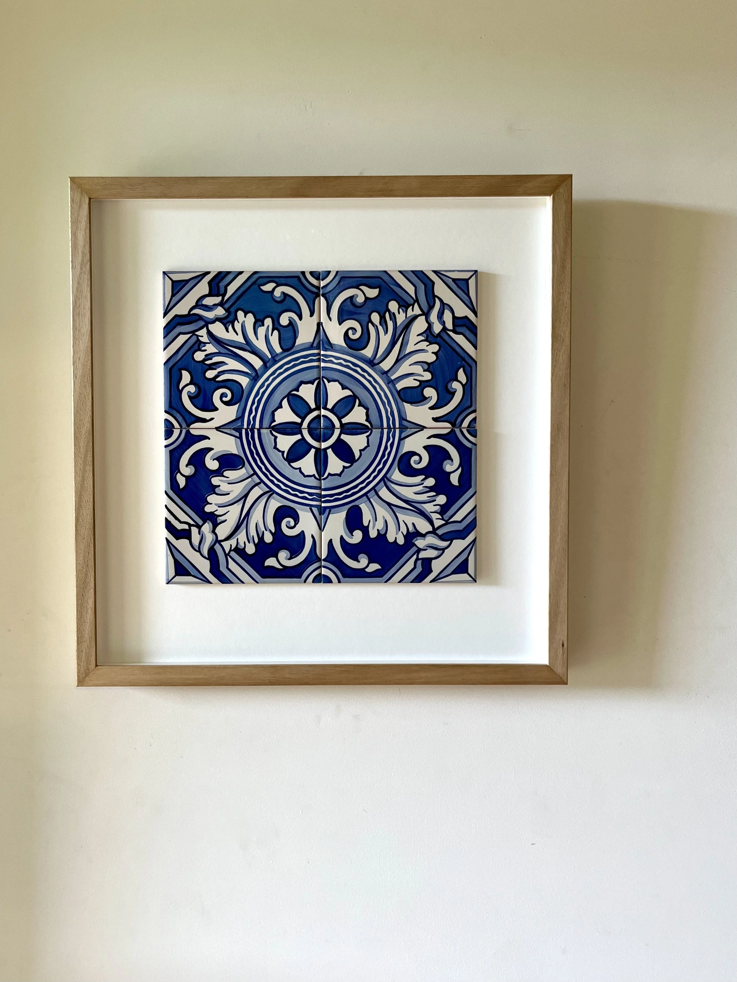 A framed artwork of blue and white ceramic tiles from Porto, Portugal with an intricate floral and geometric pattern, hanging on a plain off-white wall.